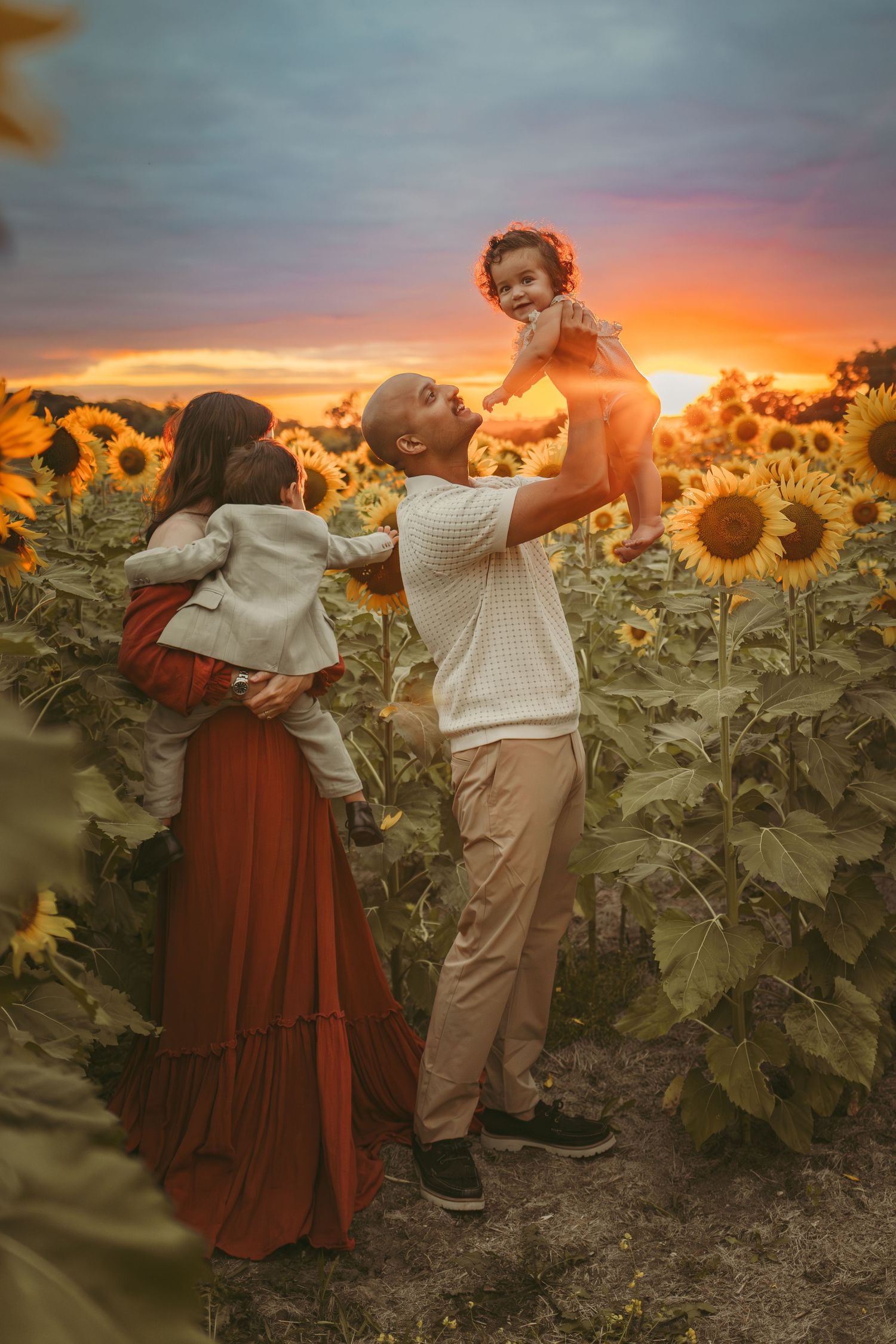 Dad holds baby in the air while mom holds brother in a Des Moines area sunflower field at sunset during a lifestyle family photo session.