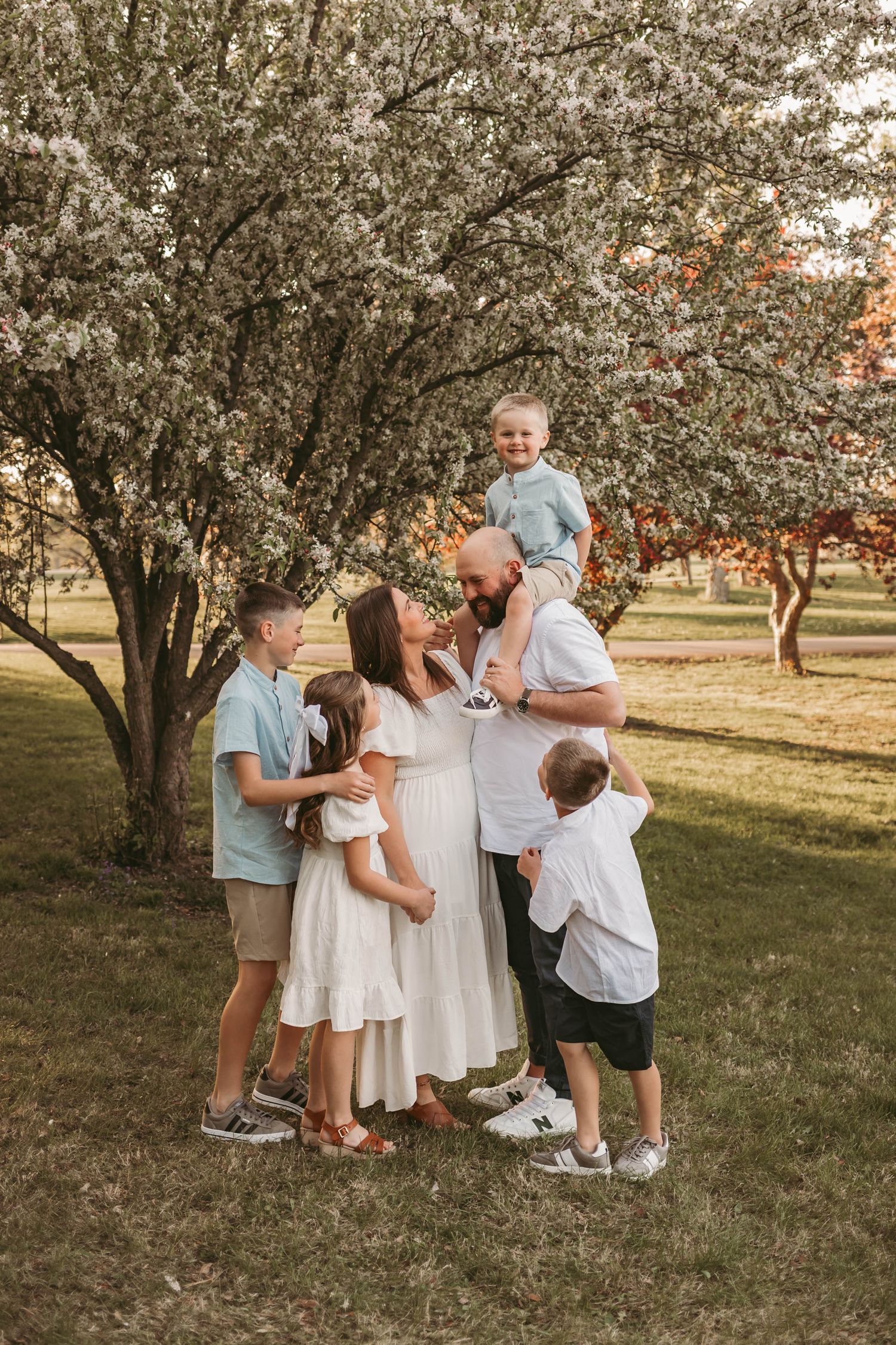 Family embraces while child sits on dad's shoulders among blossoms in a Des Moines area park during a lifestyle family photo session.