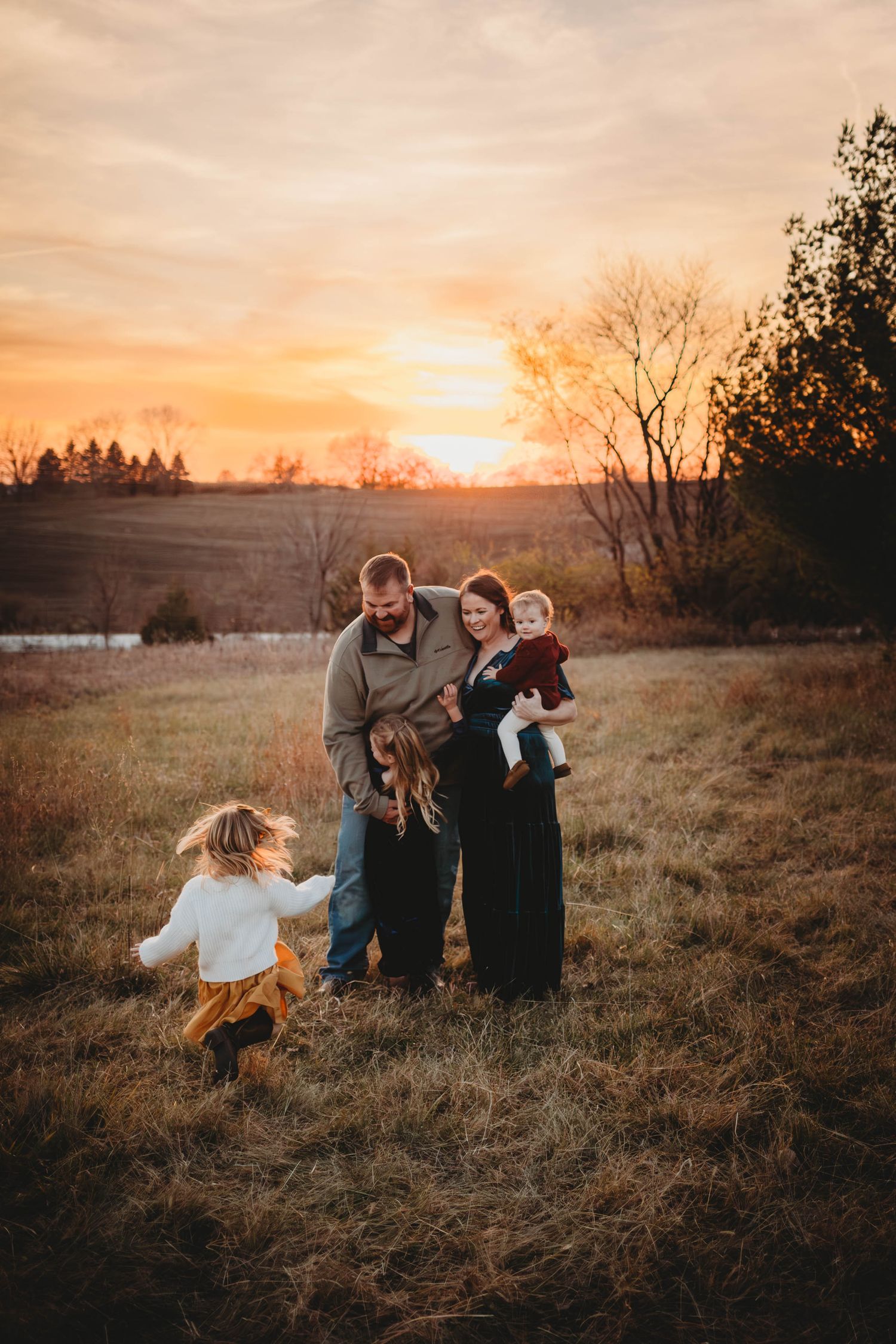 Young girls runs to embracing family's arms in a Des Moines area field at sunset during a lifestyle family photo session.
