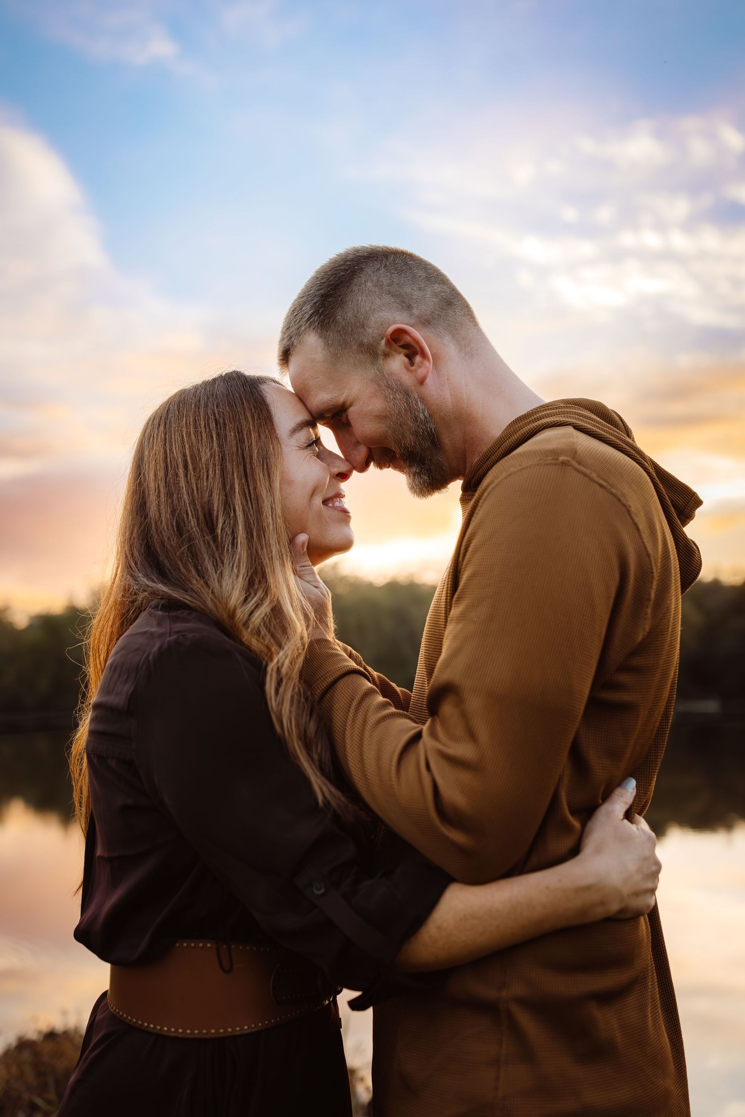 Couple hugs in front of a lake in a Des Moines area park at sunset during a lifestyle family photo session.