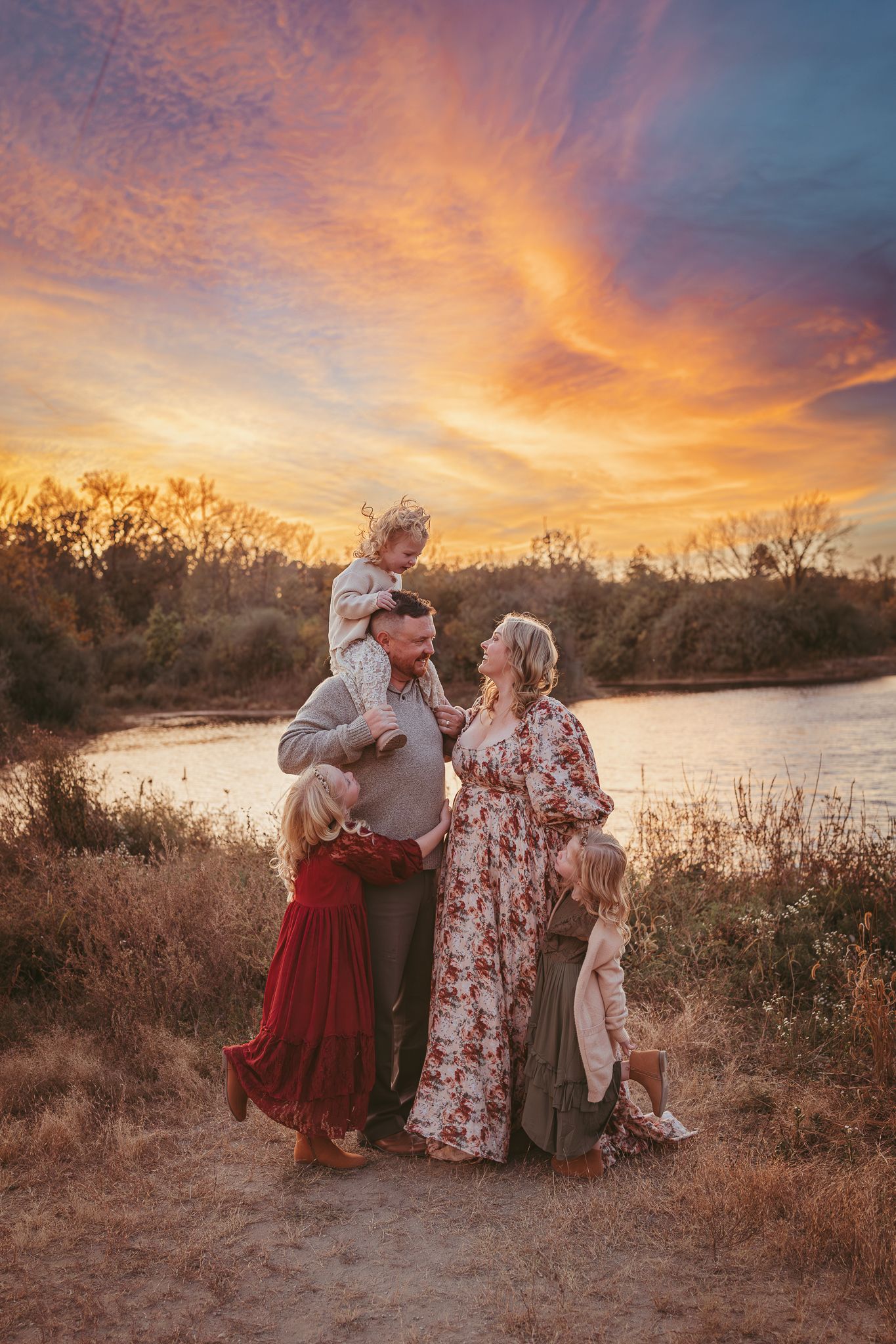 Family embraces while child sits on dad's shoulders in a Des Moines area park at sunset during a lifestyle family photo session.