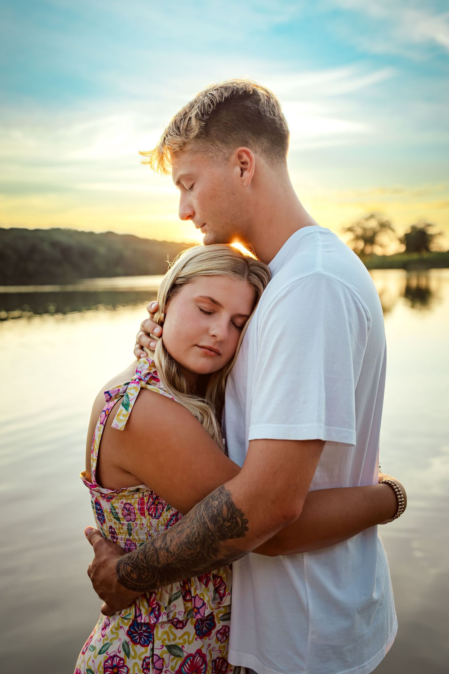 Couple hugs in front of a lake in a Des Moines area park at sunset during a lifestyle family photo session.