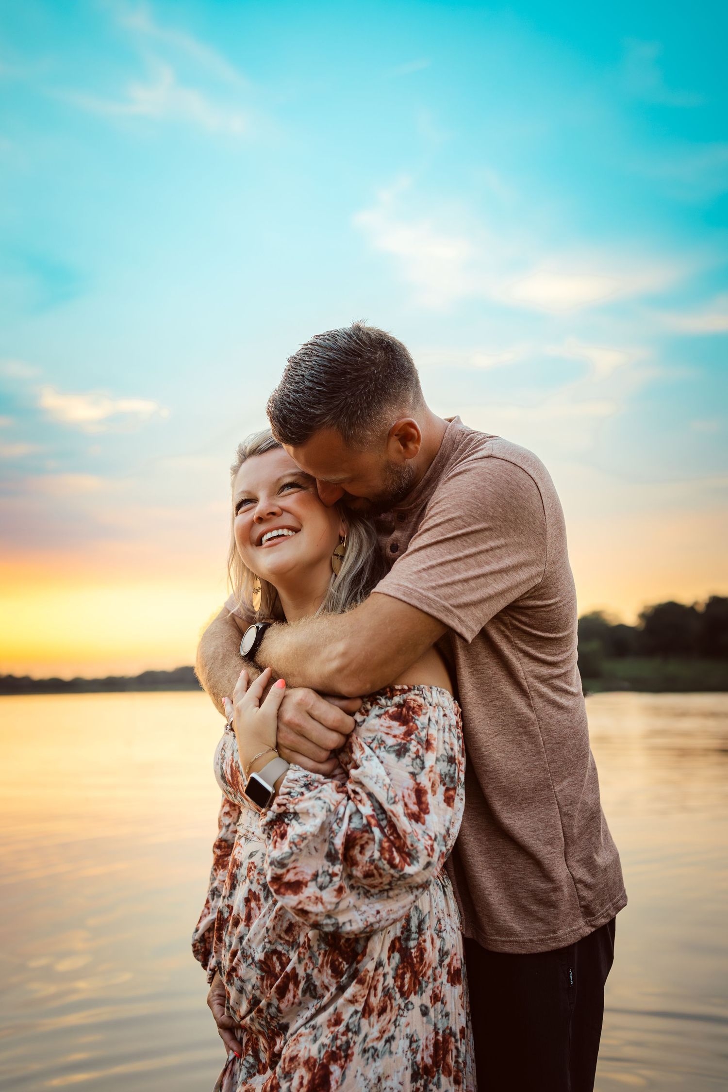 Couple hugs in front of a lake in a Des Moines area park at sunset during a lifestyle family photo session.