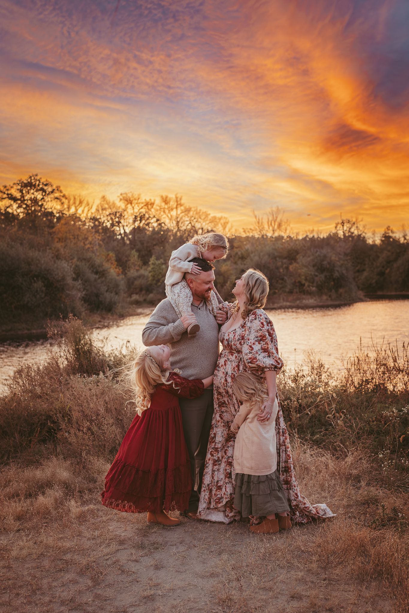 Family embraces while child sits on dad's shoulders in a Des Moines area park at sunset during a lifestyle family photo session.