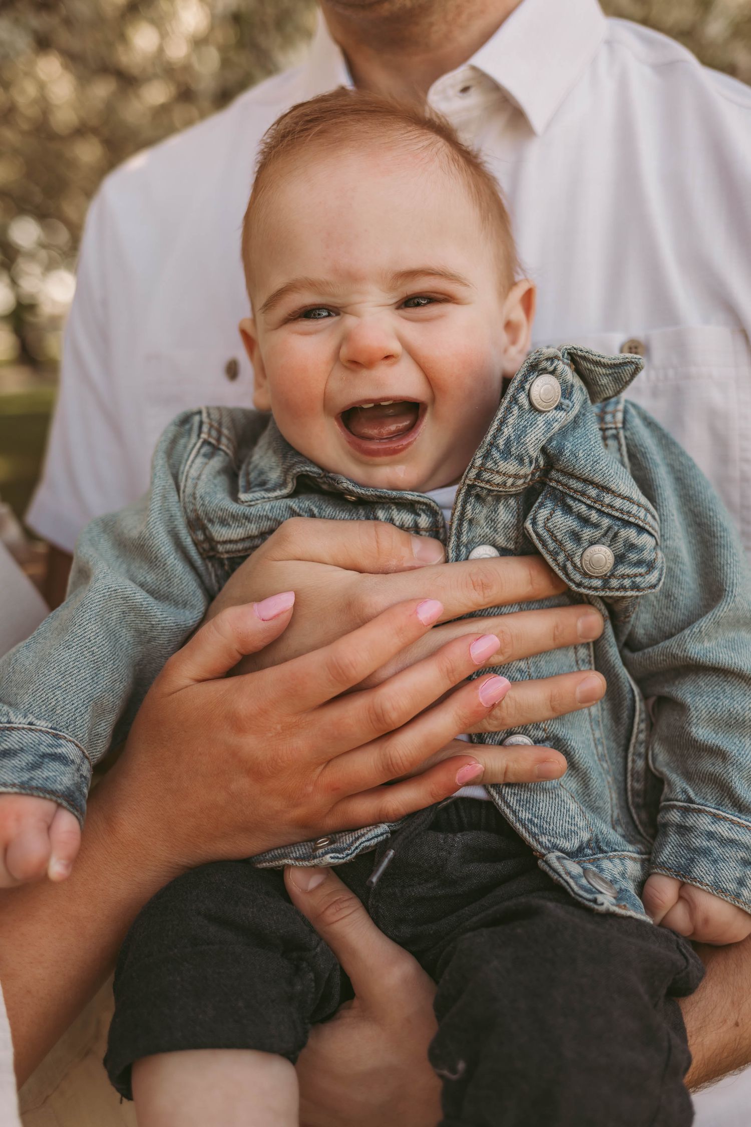 Baby boy smiles while parents hold him in a Des Moines area park during a lifestyle family child milestone photo session.