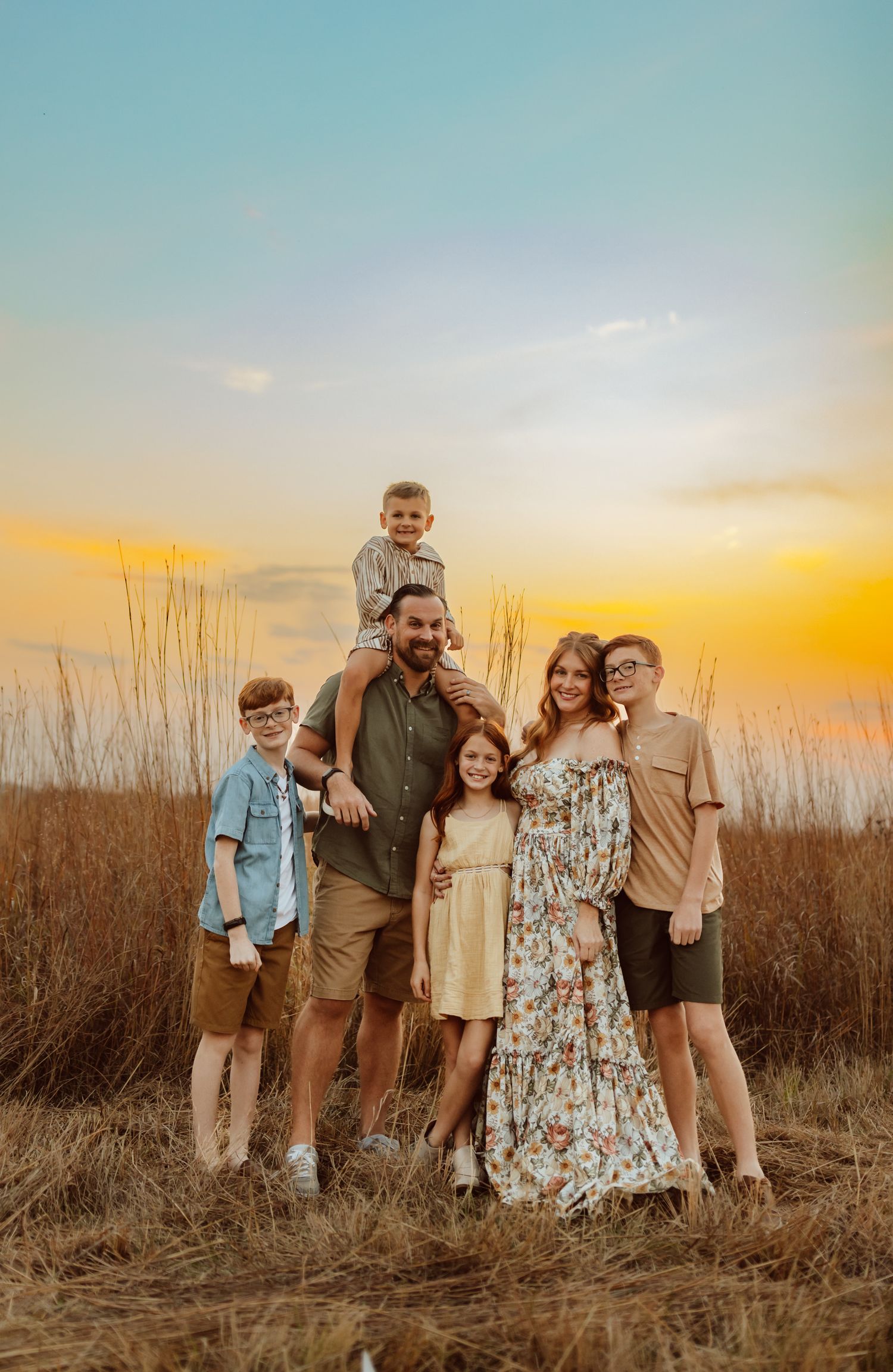 Family embraces while child sits on dad's shoulders in a Des Moines area park at sunset during a lifestyle family photo session.