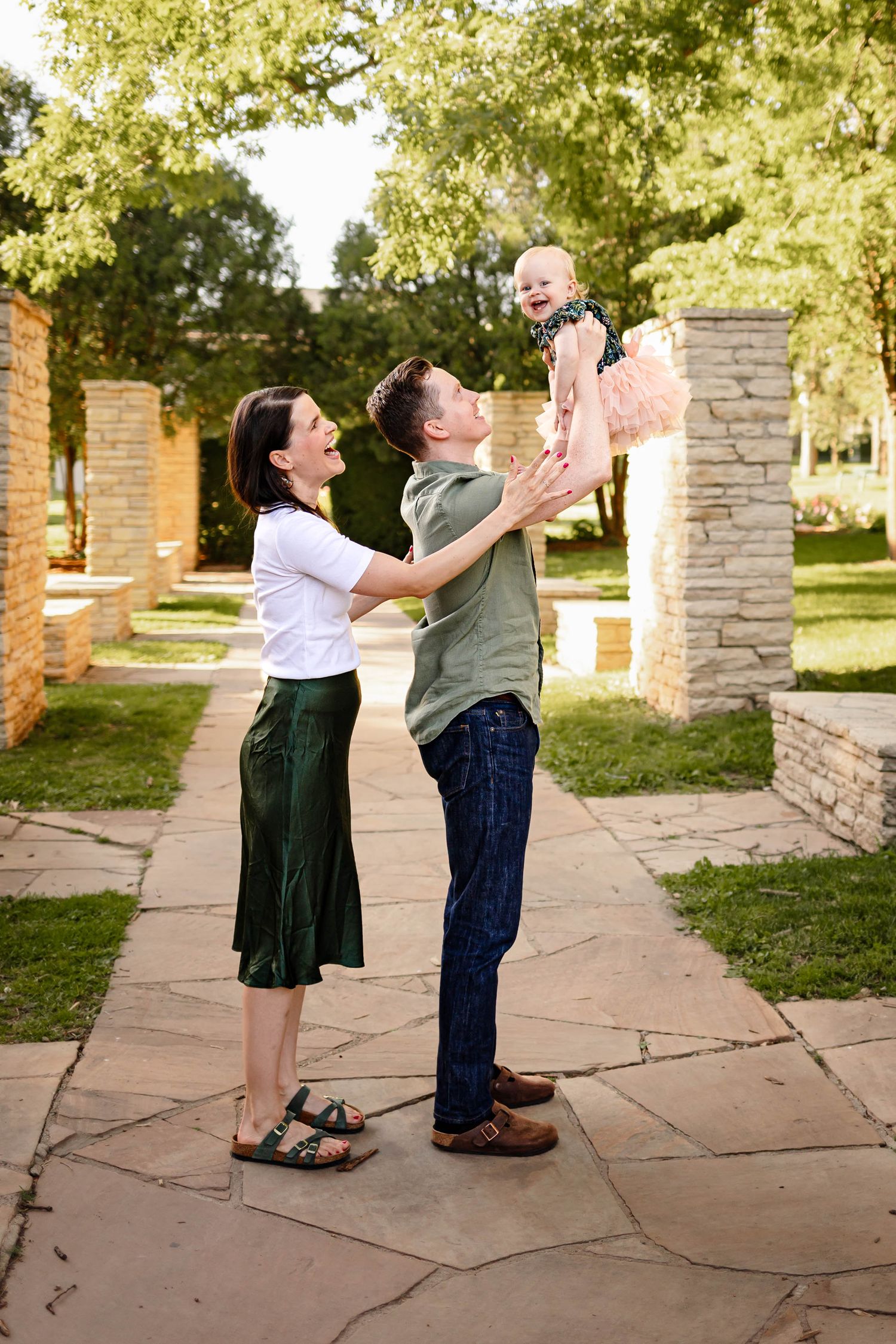 Father hold baby girl in the air as mom looks on at the Rose Garden in Des Moines during a lifestyle family photo session.
