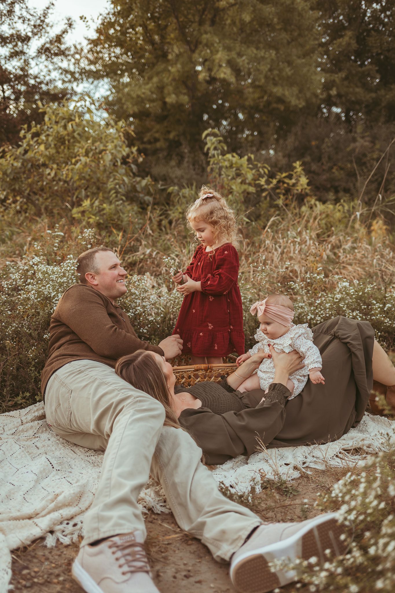 Father smiles at young daughter while mom plays with her baby in a Des Moines area park at sunset during a lifestyle family photo session.