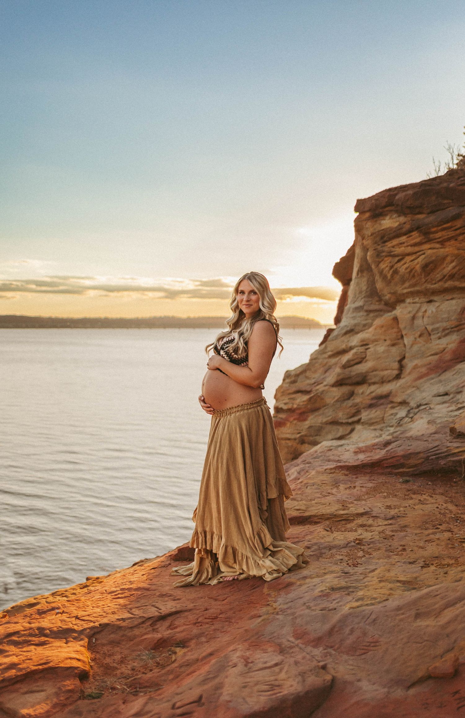 Pregnant woman stands on cliff in front of a lake in a Des Moines area park during a lifestyle maternity photo session.