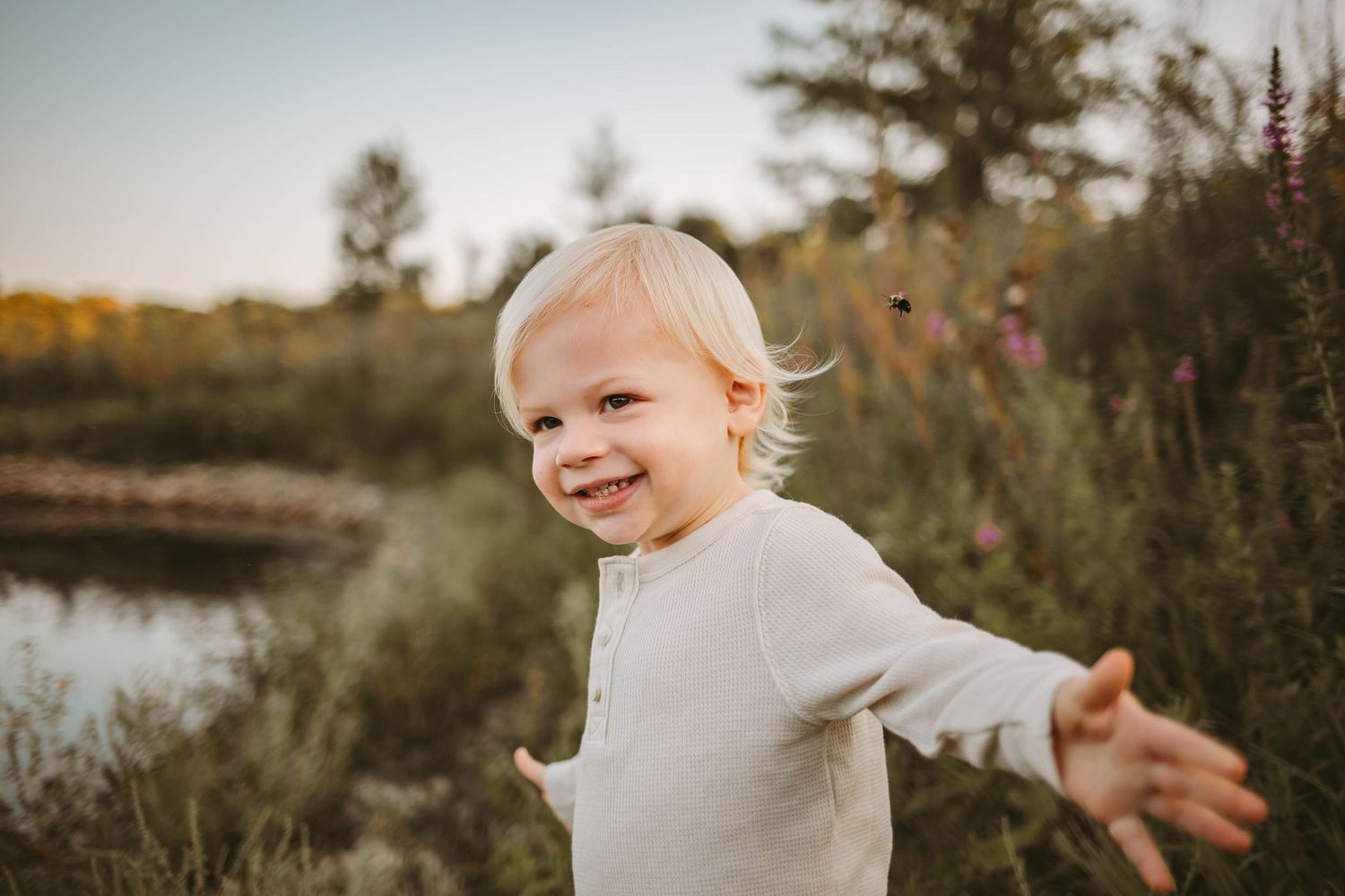 Toddler boy stands in a field in front of a lake in a Des Moines area park at sunset during a lifestyle family child milestone photo session