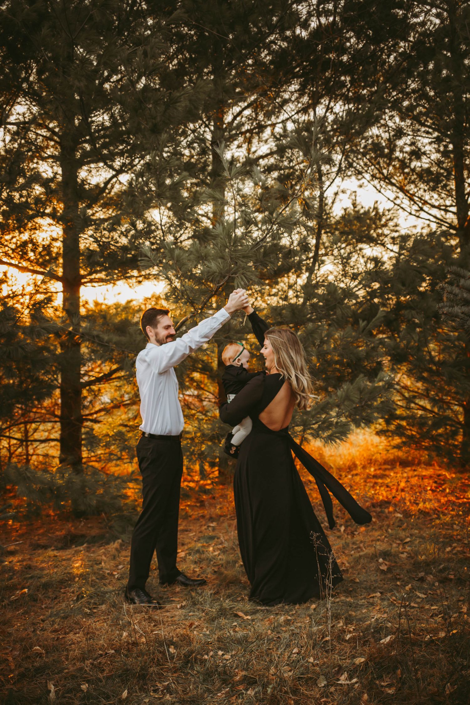 Couple dances with baby in a Des Moines area evergreen field at sunset during a lifestyle family photo session.