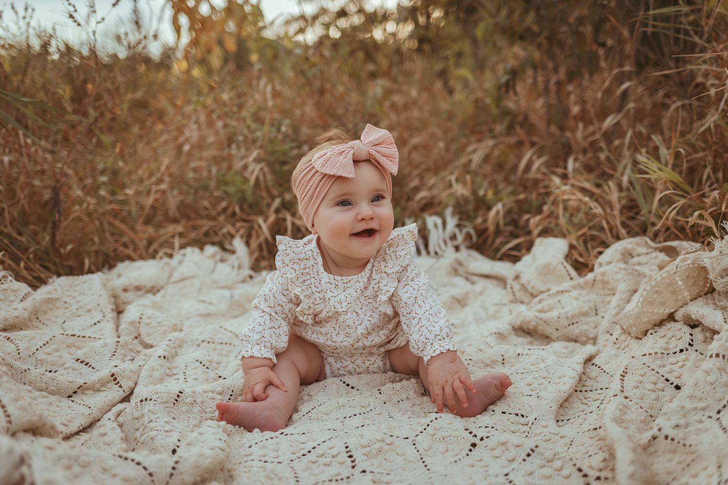 Baby girl sits and smiles on a vintage blanket in a Des Moines area park during a lifestyle family child milestone photo session.