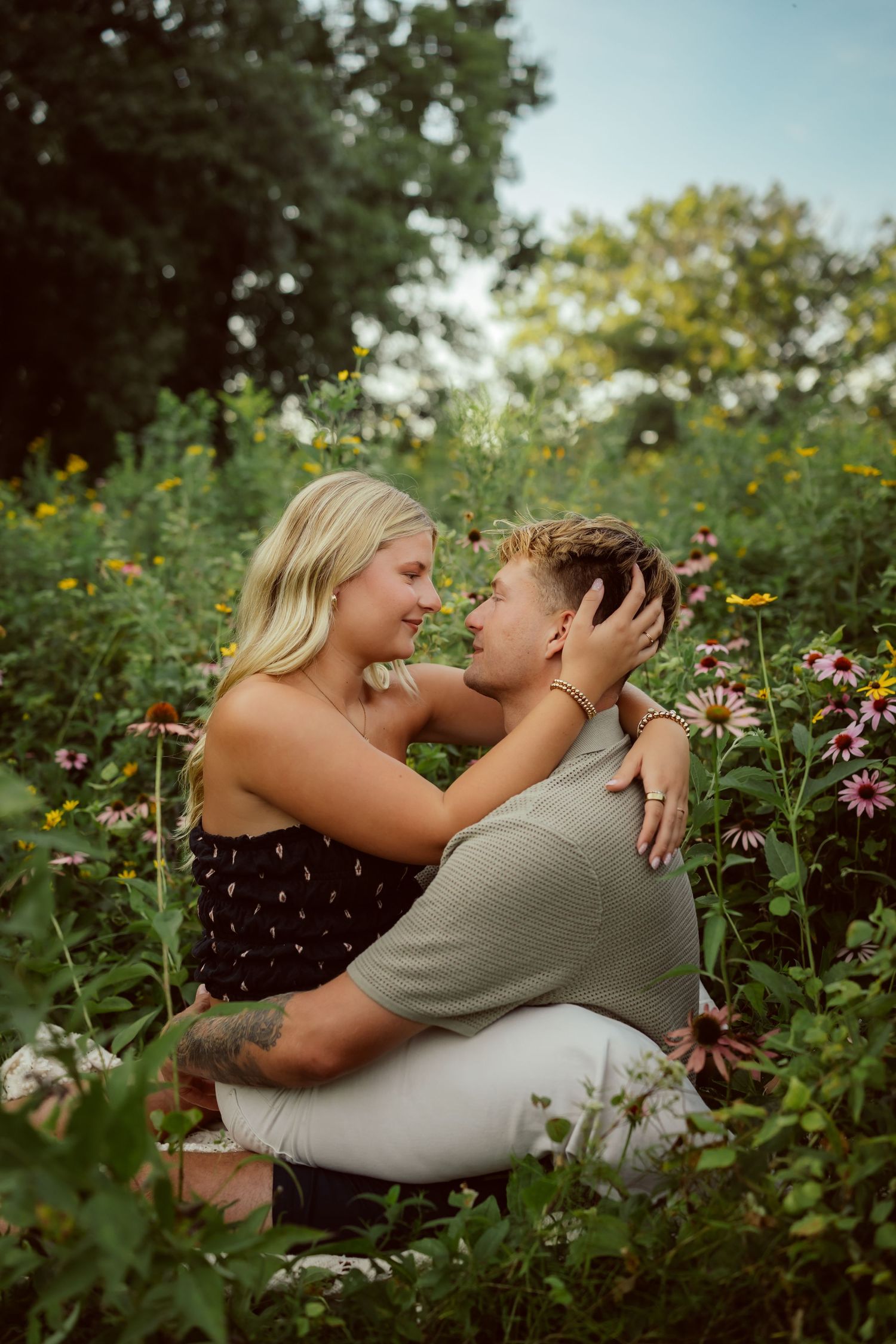 Couple sits looking in each others eyes in a field of flowers in a Des Moines area park at sunset during a lifestyle family photo session.