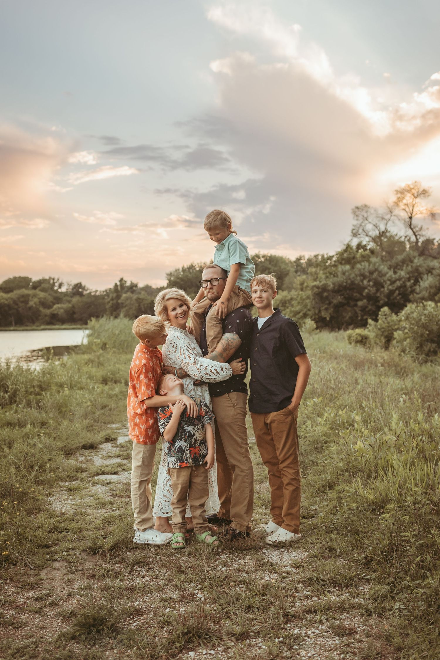 Family embraces while child sits on dad's shoulders in a Des Moines area park at sunset during a lifestyle family photo session.