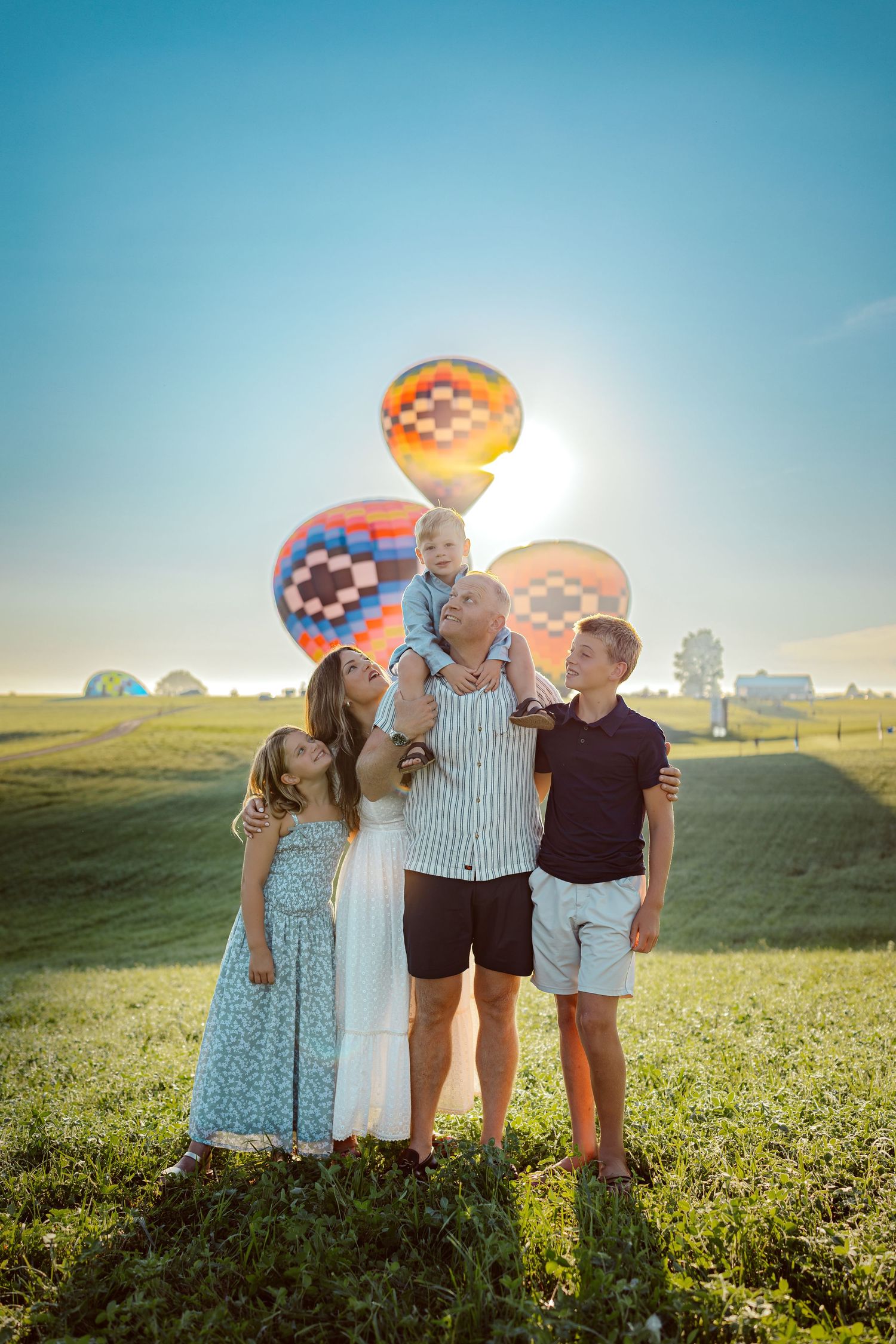 Family stands before hot air balloons in a Des Moines area park at sunrise during a lifestyle family photo session.