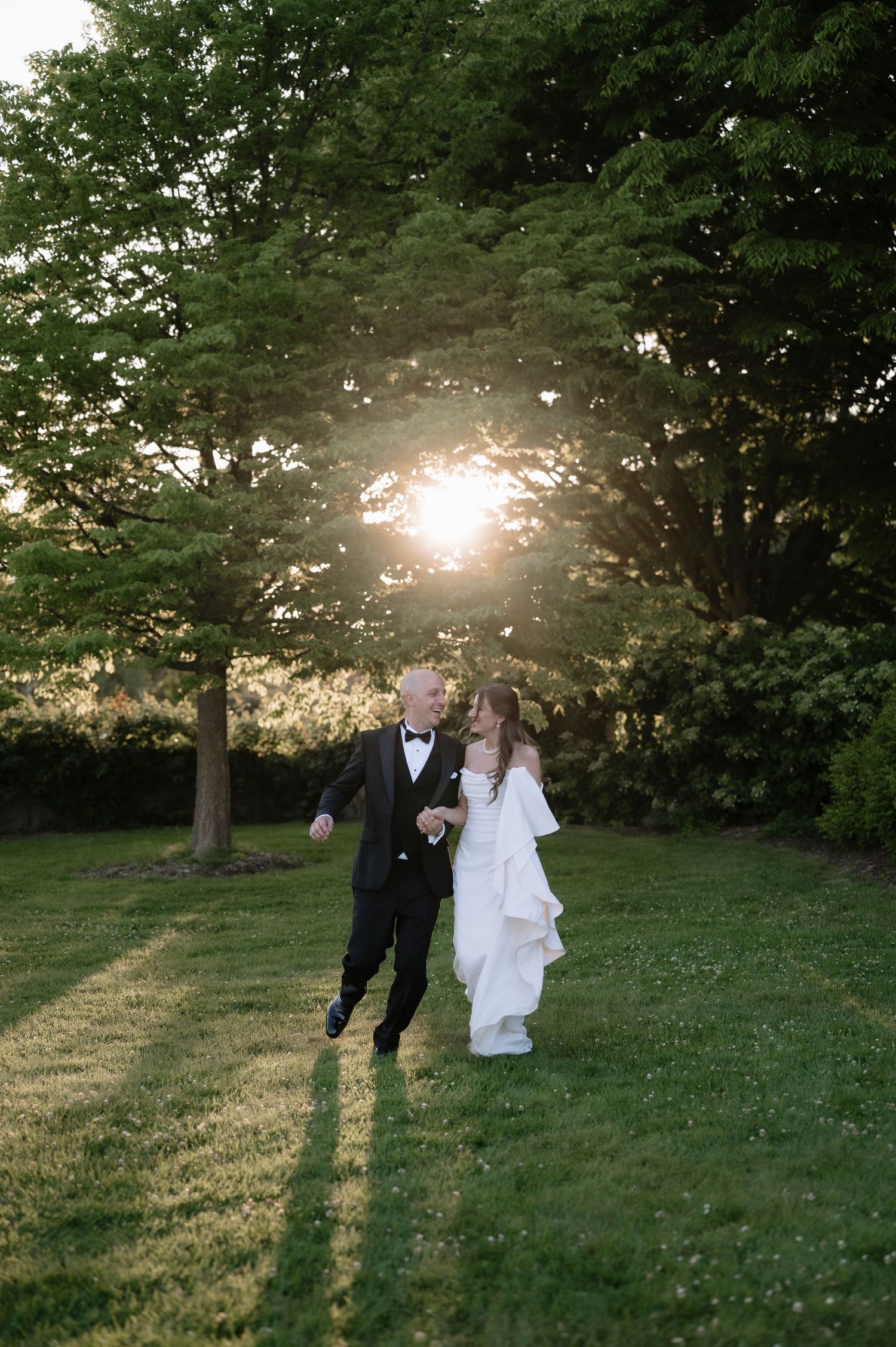 A couple in wedding attire walk together on a grassy lawn at sunset with sunlight filtering through trees.