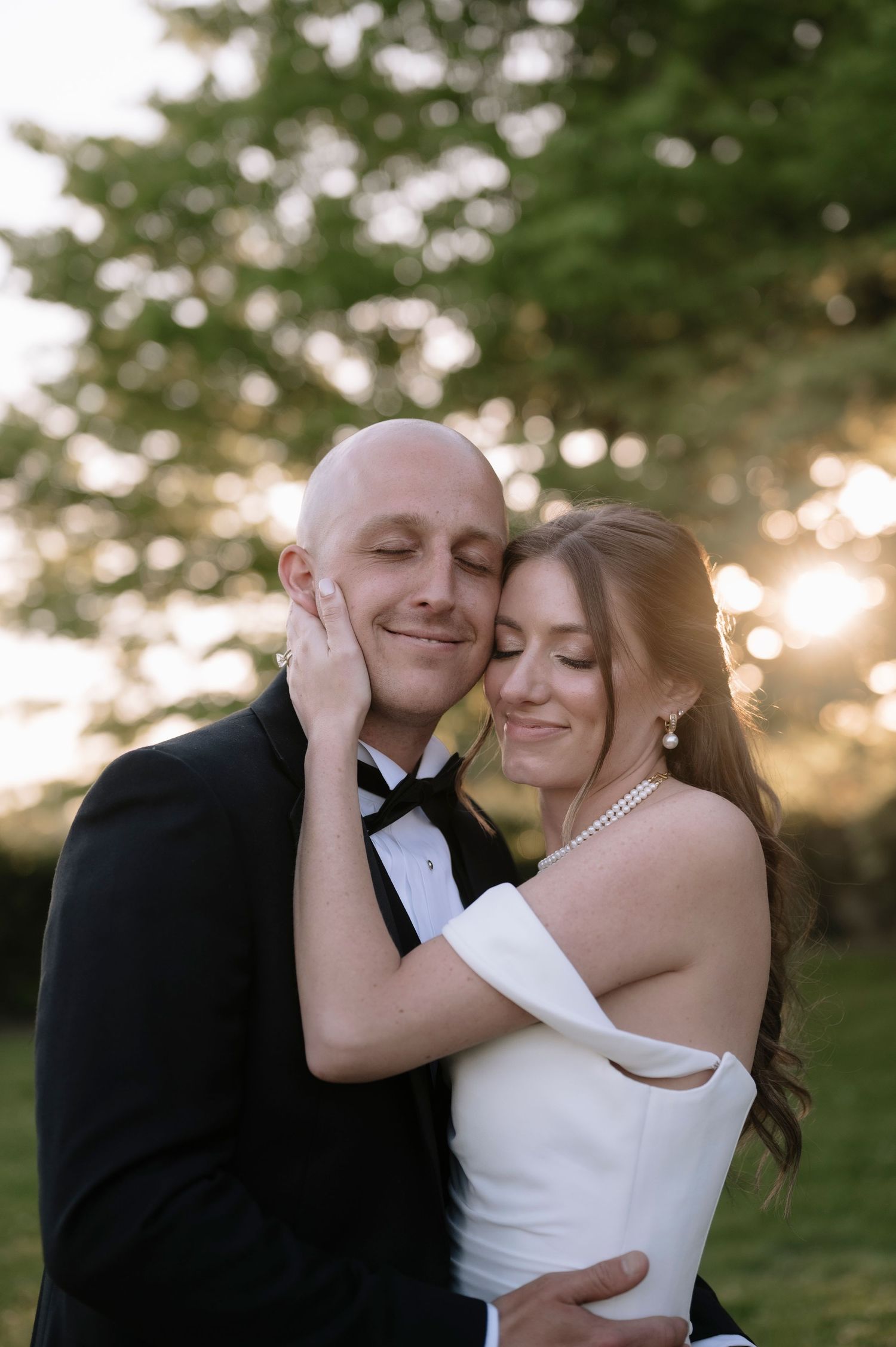 A loving couple shares an intimate moment outdoors at sunset in formal wedding attire with a blurred nature backdrop.