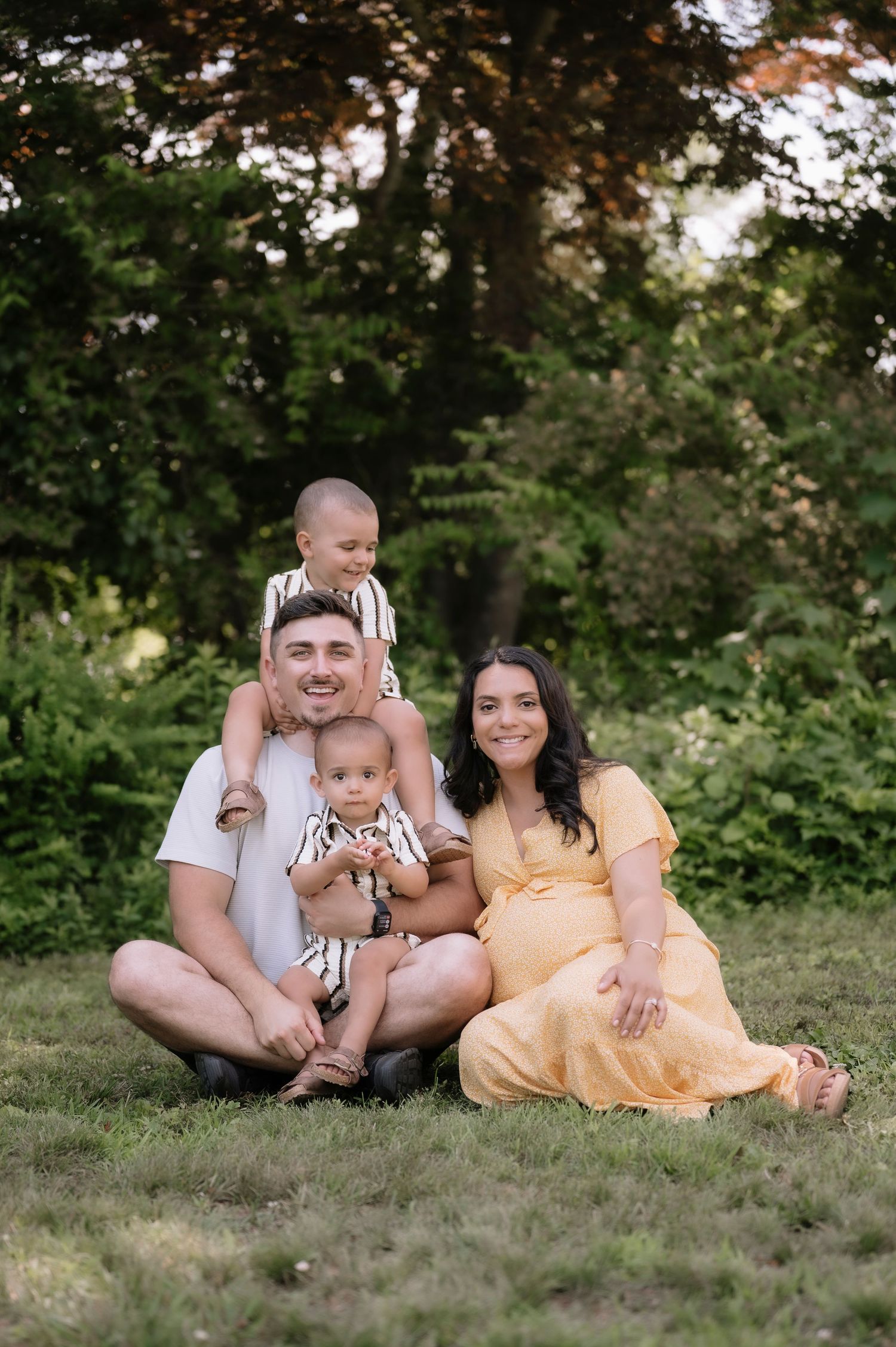A happy family of four poses outdoors on grass, with a pregnant mother in a yellow dress and father holding two young children.