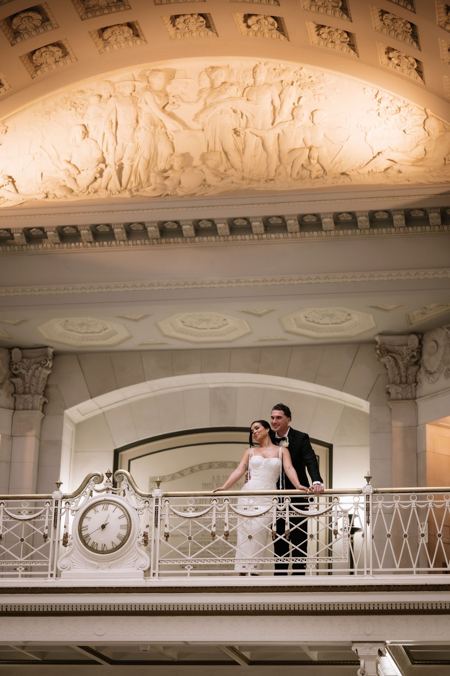 Bride and groom stand on an ornate balcony with a clock beneath elegant relief sculptures in a grand venue.