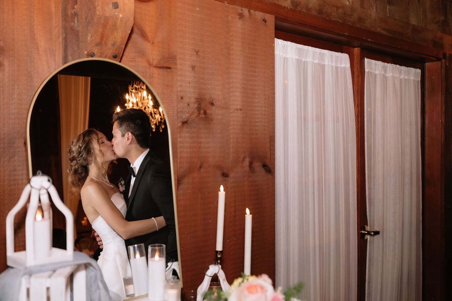 Bride and groom sharing a kiss reflected in an elegant arch mirror at their rustic wedding reception.