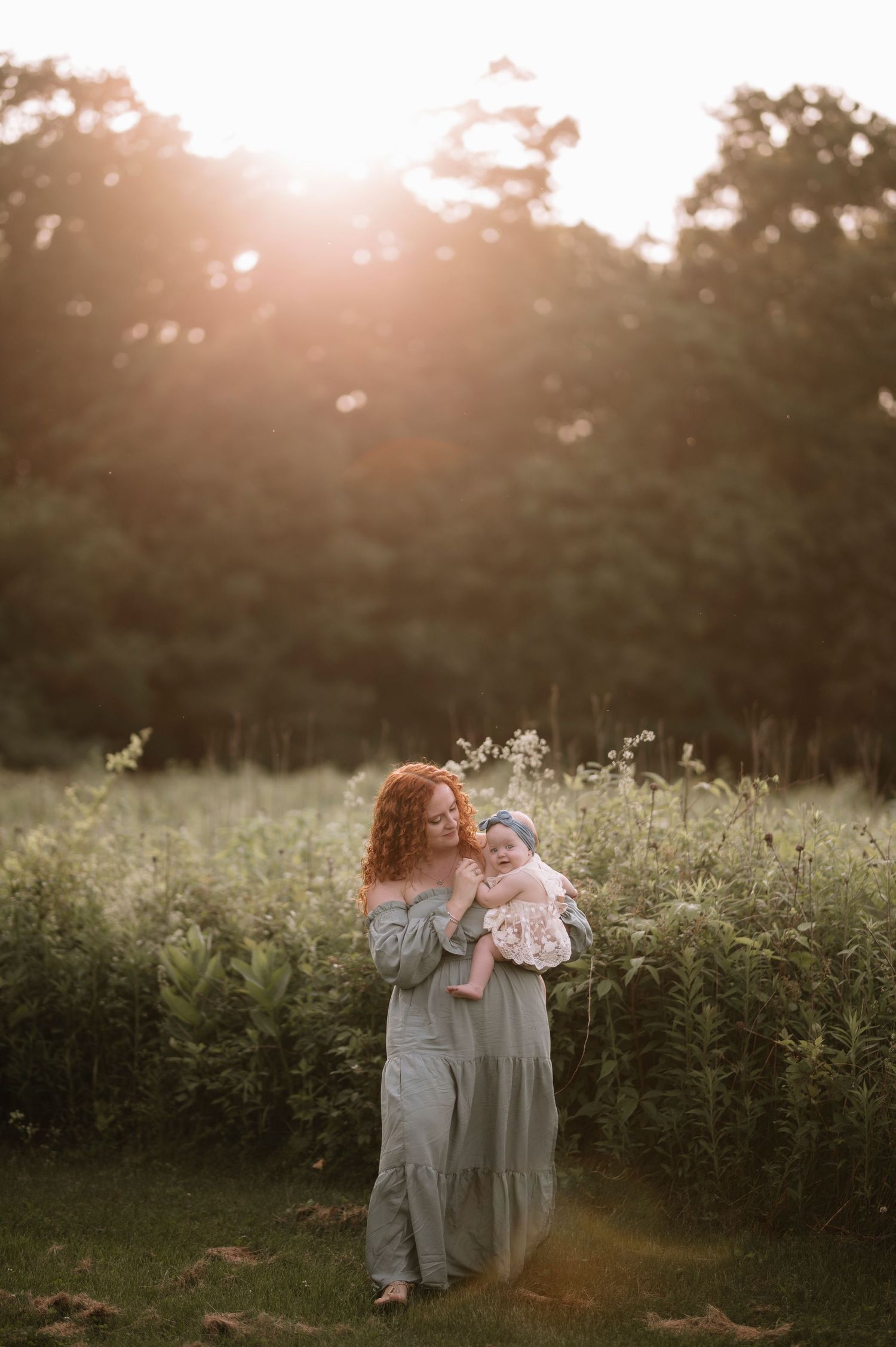 A mother and baby share a tender moment in a sunlit meadow at golden hour, surrounded by wildflowers and tall grass.