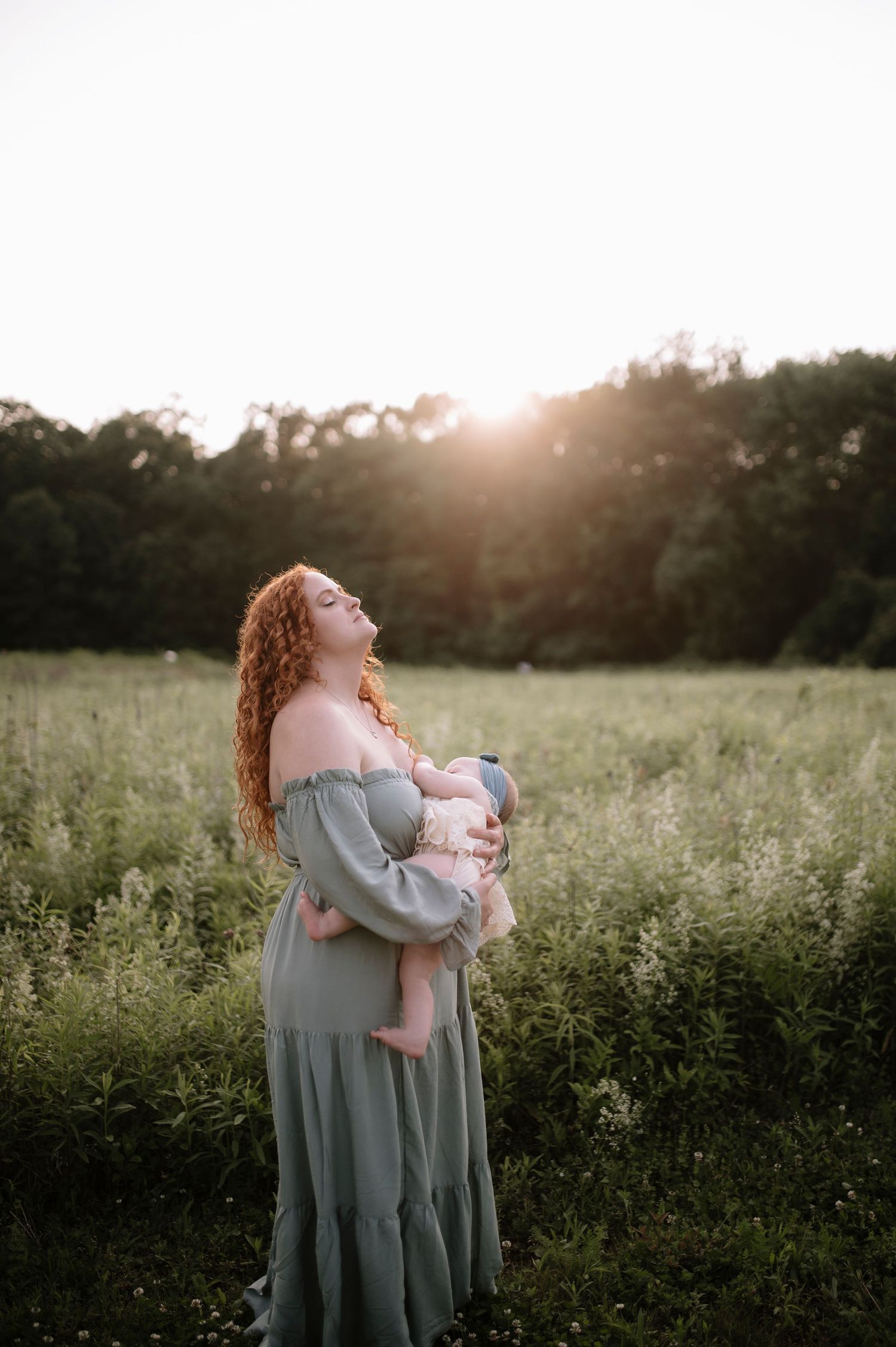Woman in flowing sage green dress holding a baby stands in a golden sunset meadow with tall grass and trees.
