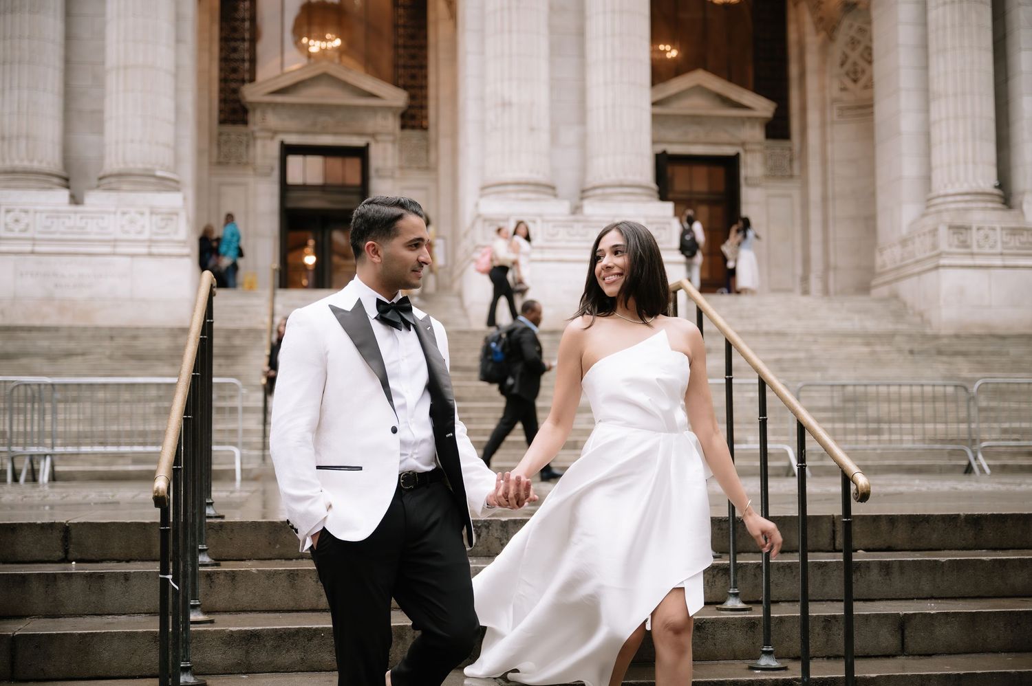 A wedding couple descends the grand stone steps of a classic building with white columns on a sunny day.