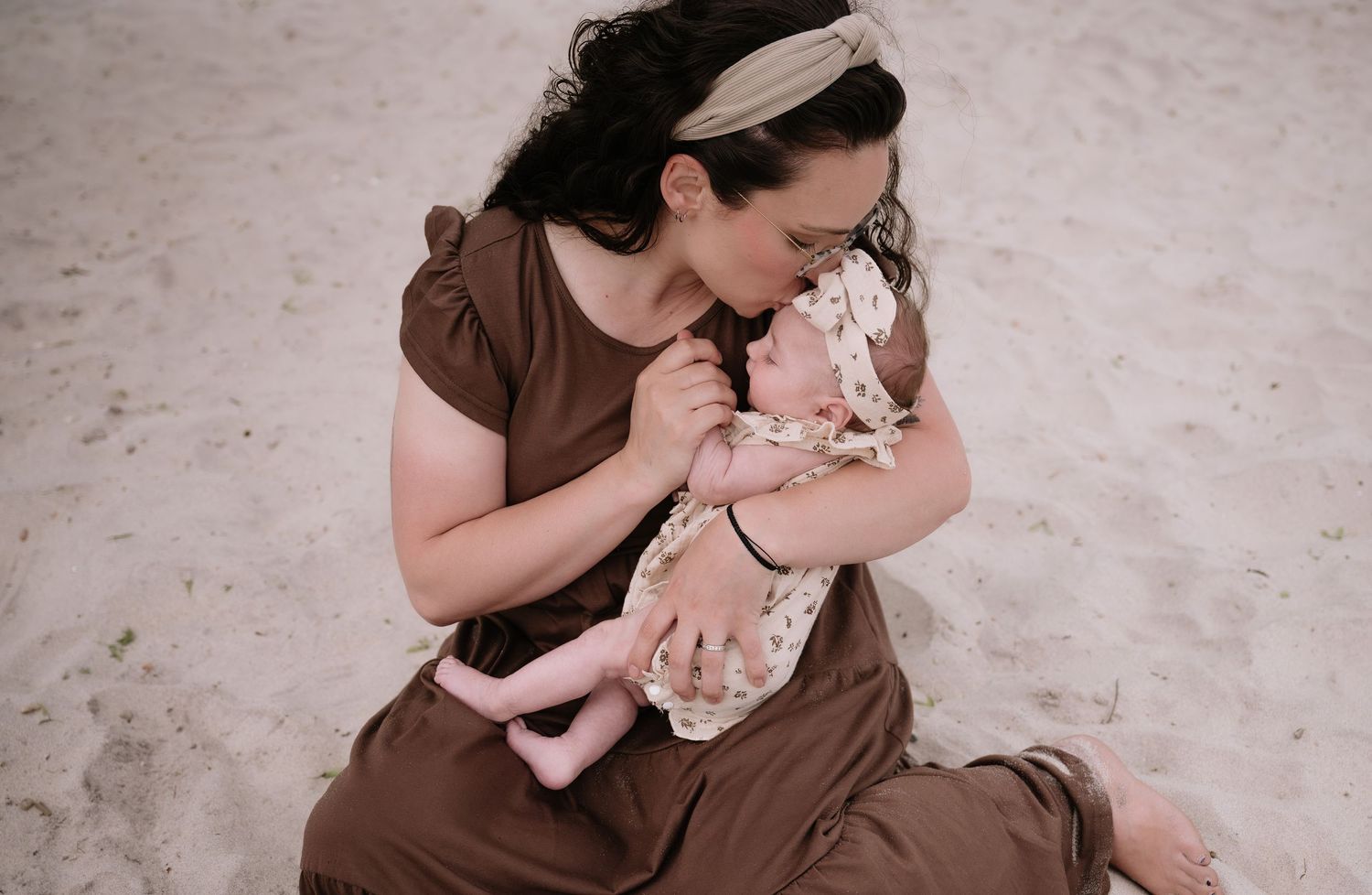 A person in a brown dress sits on the ground holding a small baby dressed in light colored clothing.