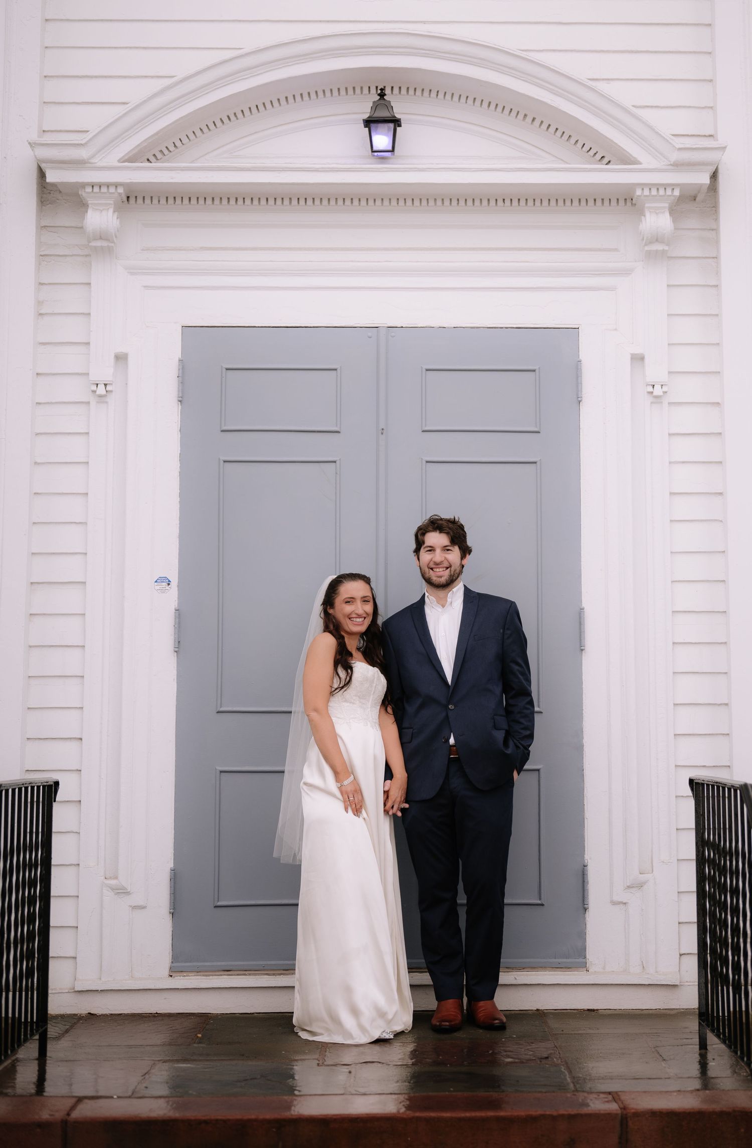 Couple in wedding attire poses in front of large gray doors beneath classic architectural archway.