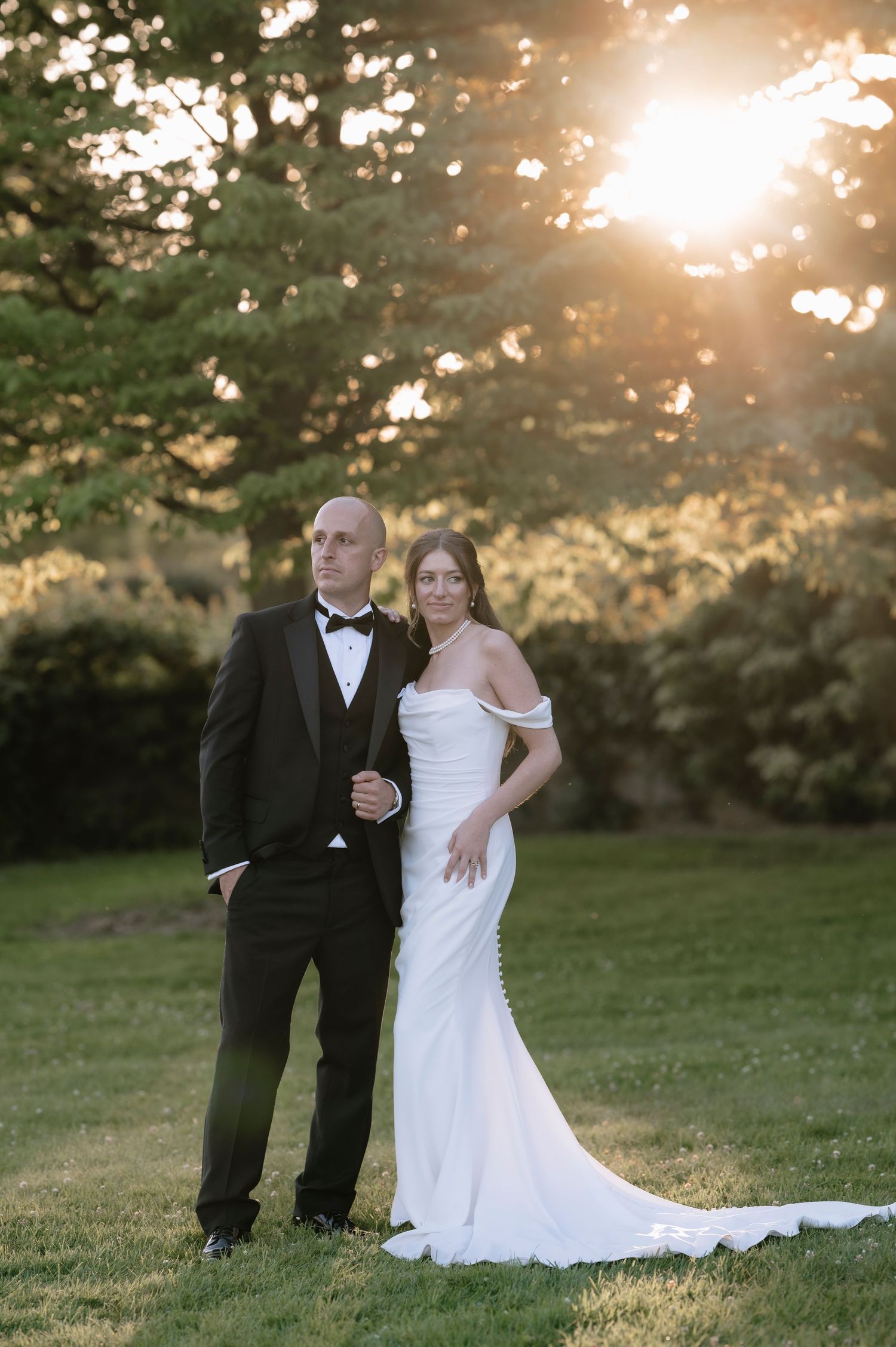 A couple poses together outdoors at sunset, with the bride wearing a white gown and groom in black tuxedo.