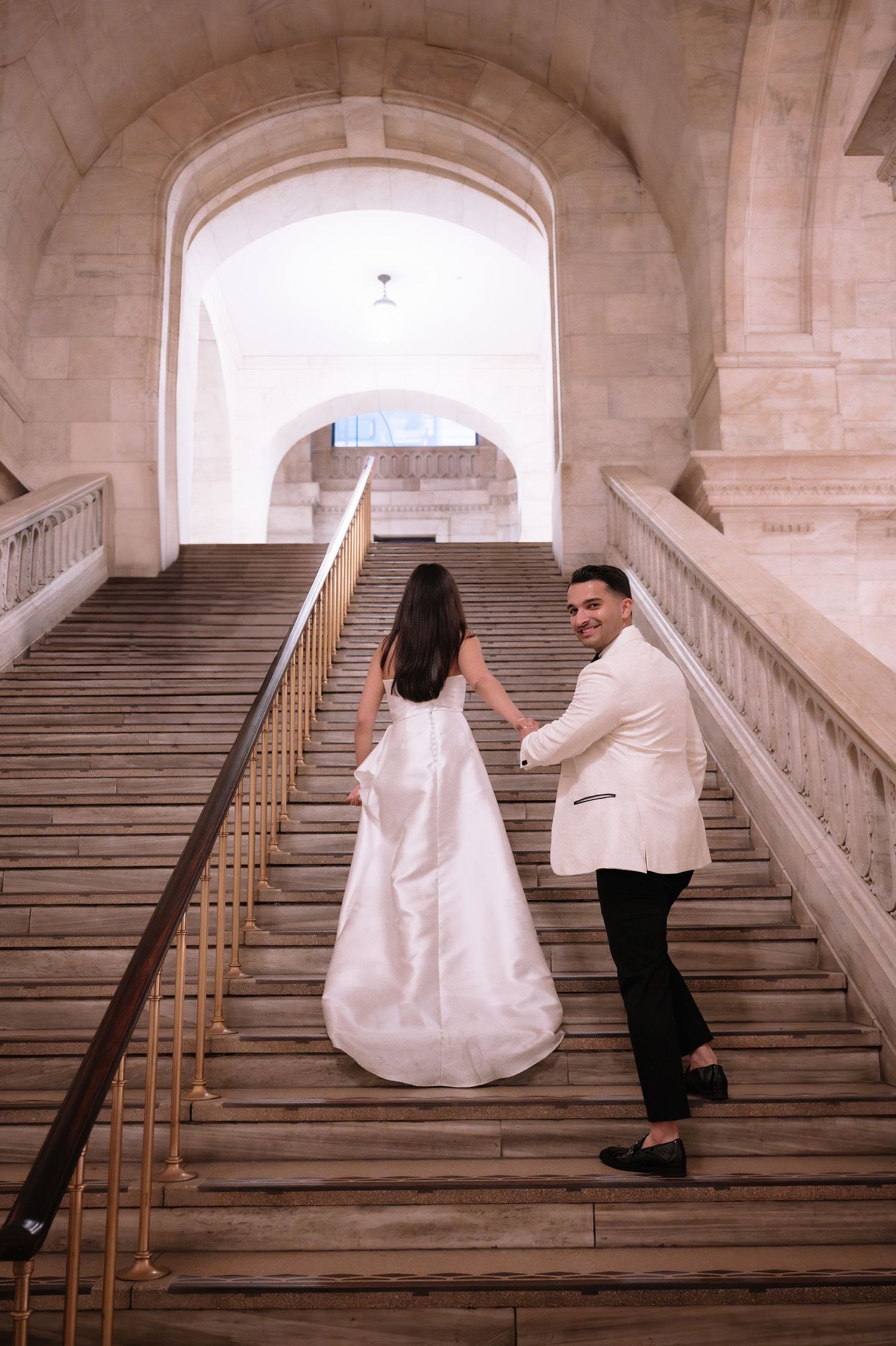 A couple in elegant white attire pose on grand marble stairs under an arched passageway.