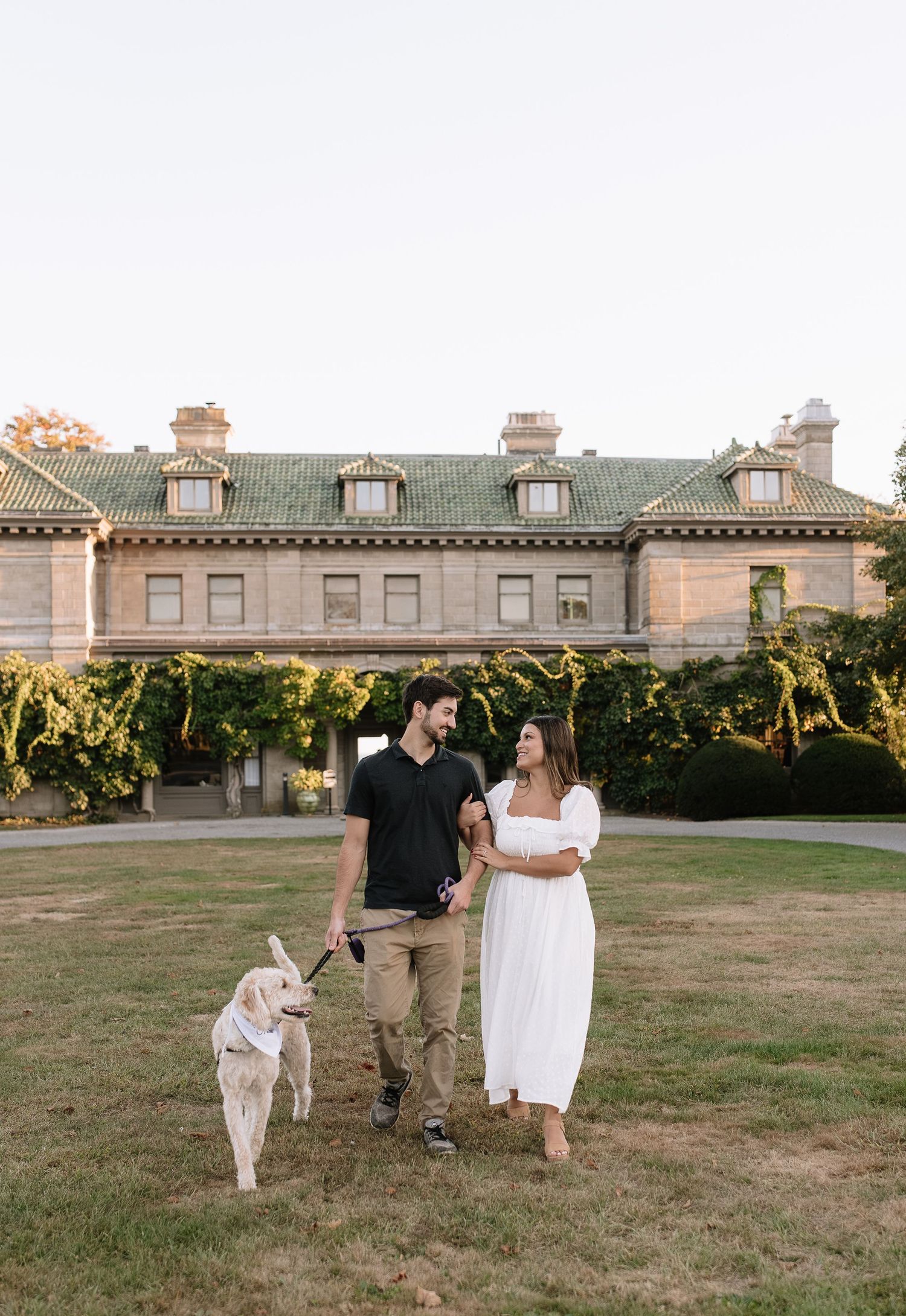 A couple walks their white dog on the lawn of a stately mansion during golden hour.
