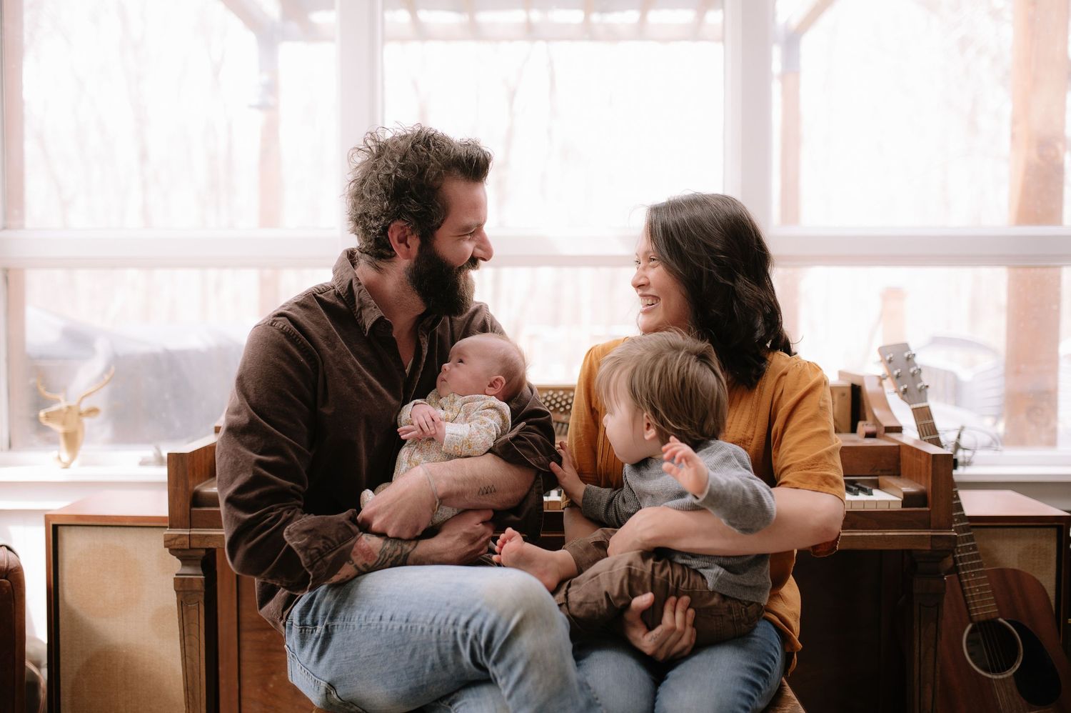 A couple sitting together on cushions lovingly holding their baby and toddler in a warm, sunlit room.