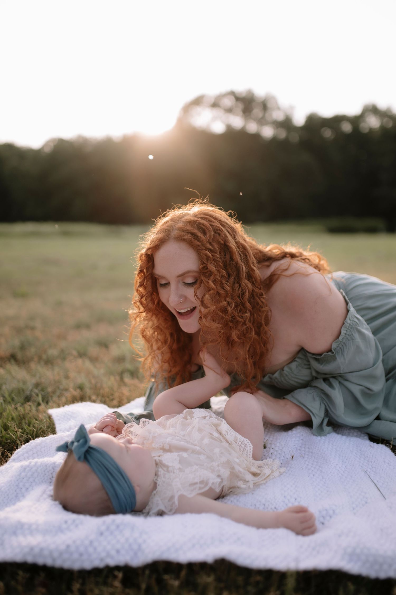 A tender moment shared between a woman with red curly hair and a baby lying on a white blanket at sunset in a grassy field.