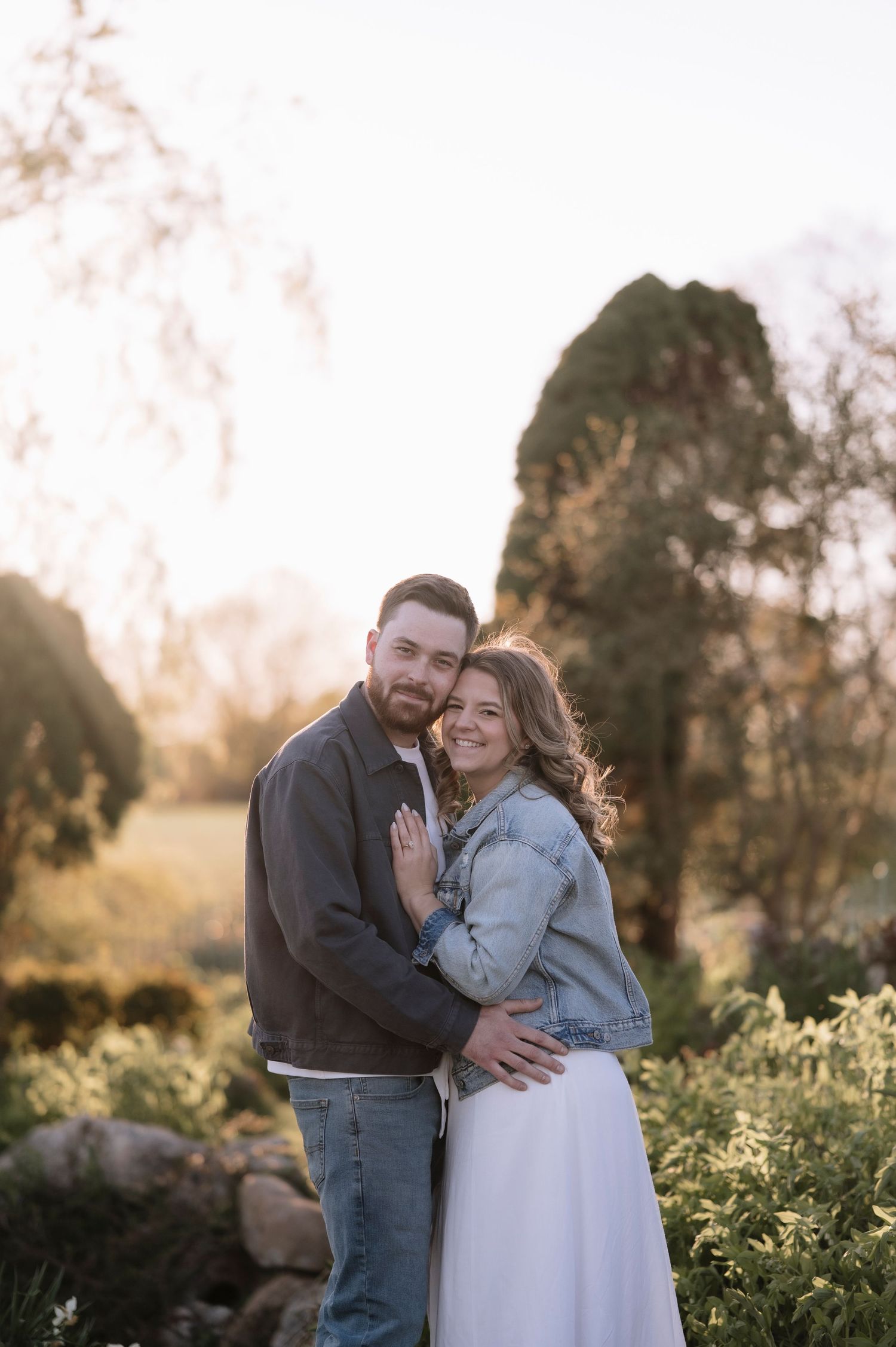 Couple embracing in a sunlit garden, woman in denim jacket and white skirt smiling at camera during golden hour.