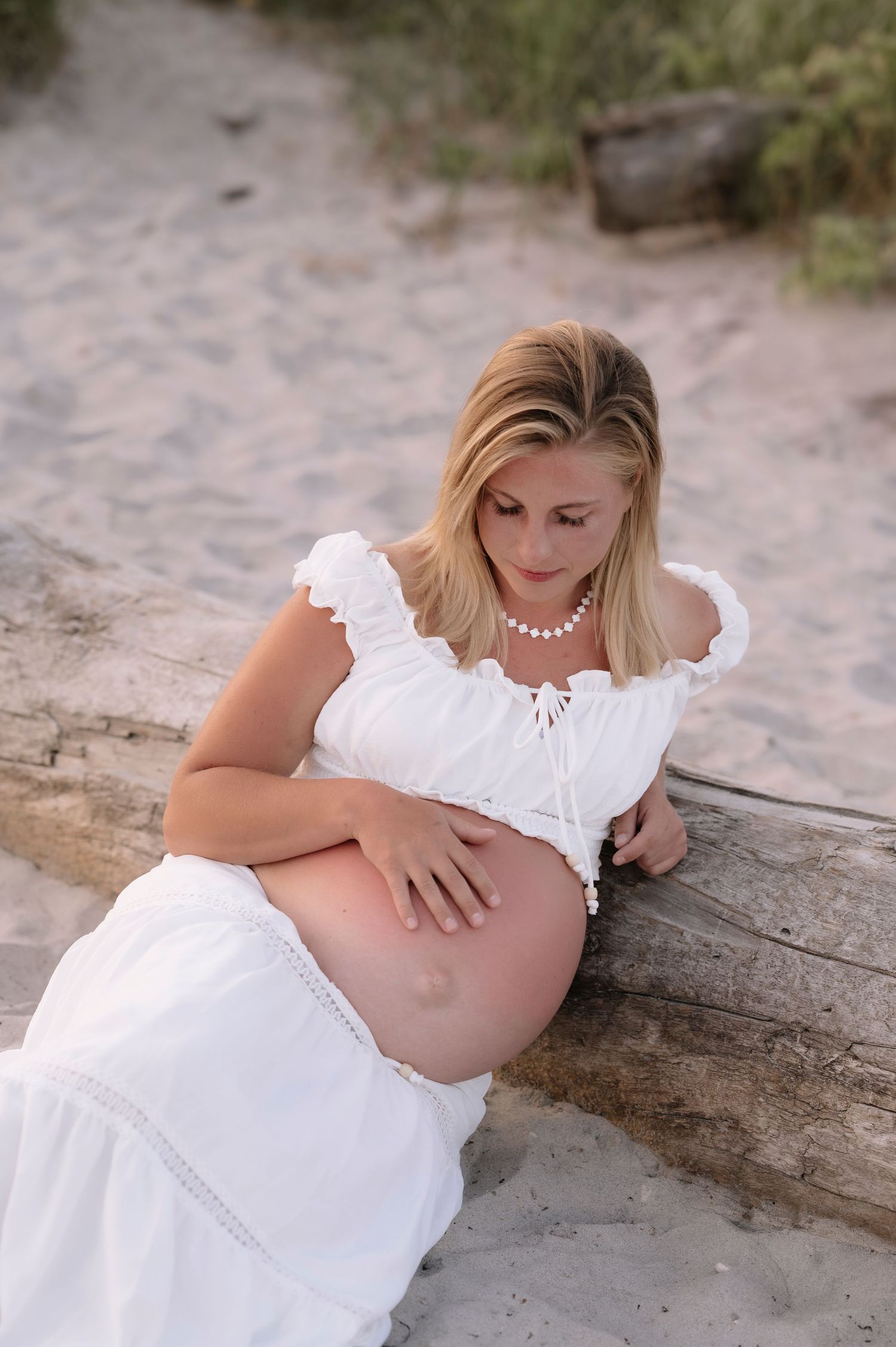 Pregnant woman in white dress sitting against driftwood on sandy beach, cradling her baby bump.