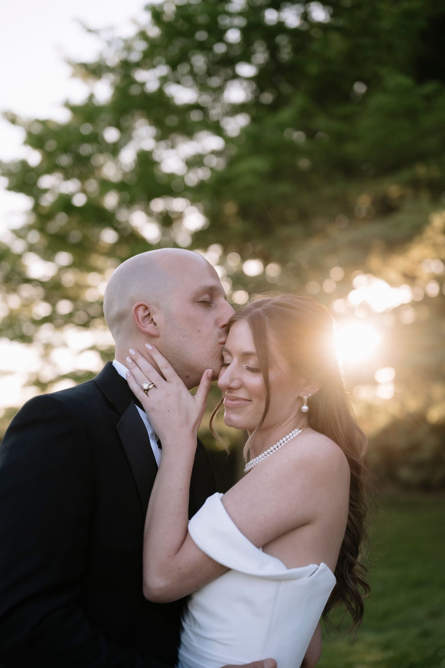 Romantic wedding photo of couple sharing intimate moment at sunset with soft bokeh through trees in background.