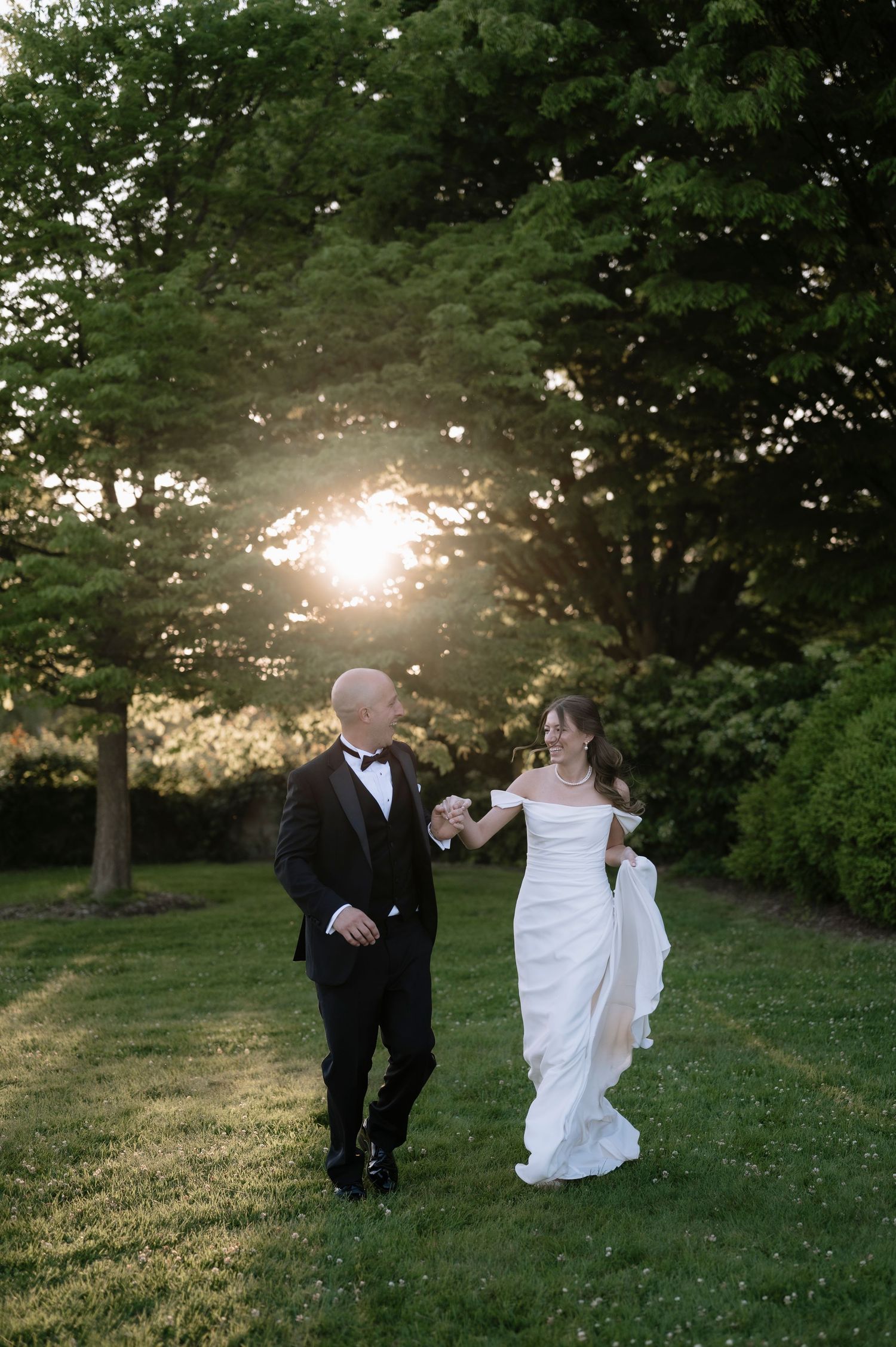 A couple in formal wedding attire walks together on a grassy lawn at sunset with trees creating a romantic backdrop.