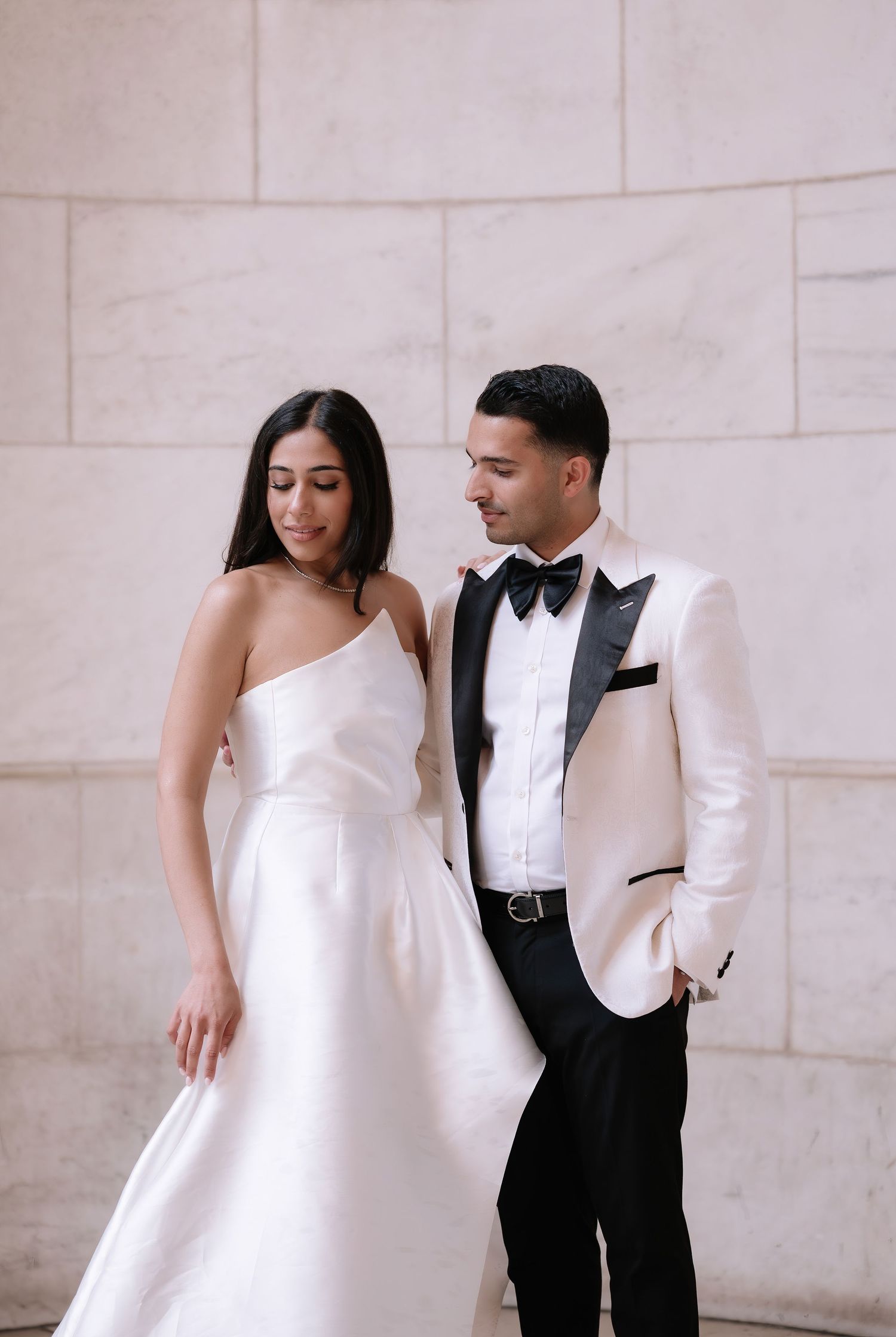 A couple in formal wedding attire poses against a marble wall, wearing a white gown and white tuxedo jacket with black pants.