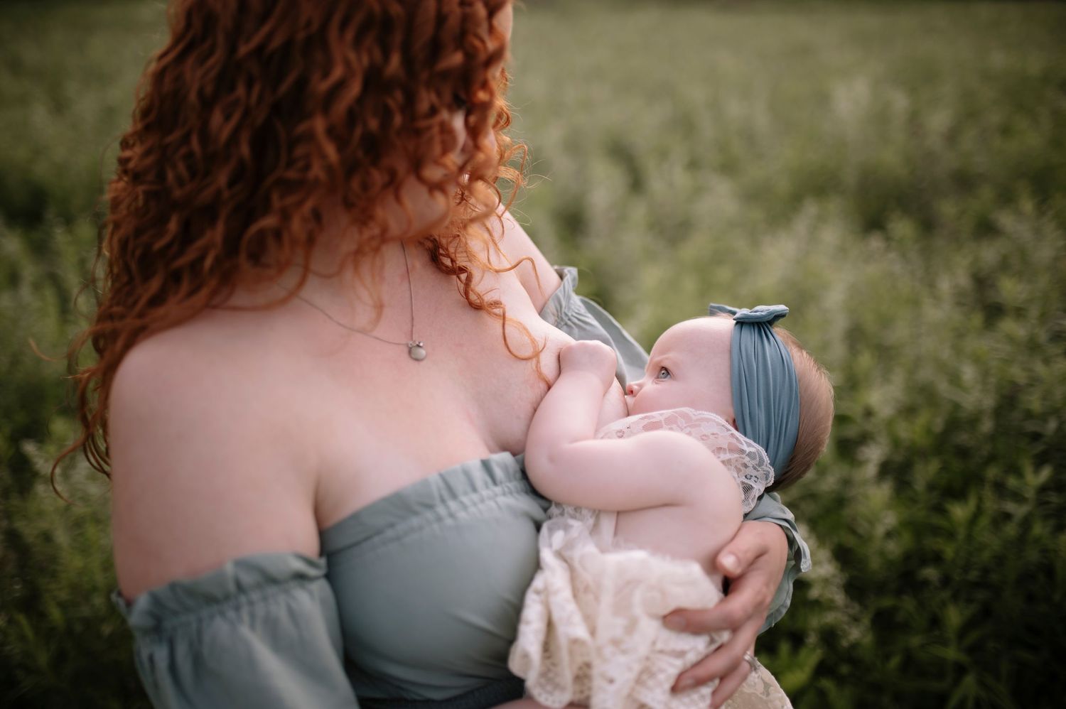 A baby nurses while being held in arms against a soft grey dress, with a natural outdoor background in soft focus.