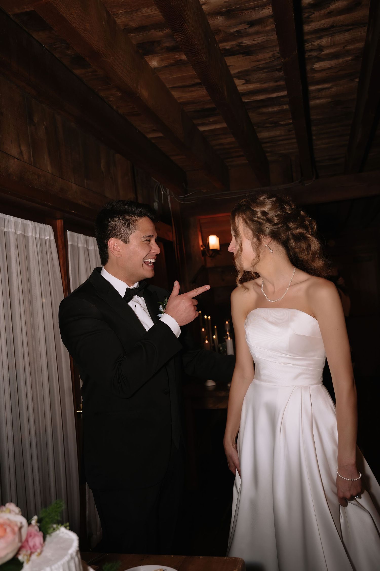 Groom in black tuxedo points at smiling bride in strapless white gown inside rustic wood-beamed wedding venue.