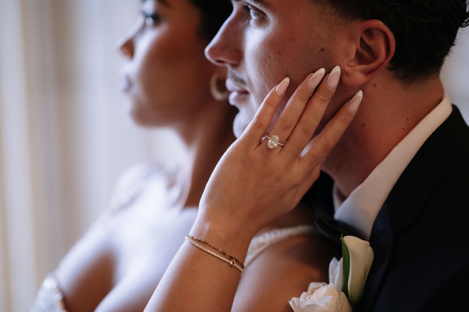 Bride tenderly touches groom's face showing elegant engagement ring and bracelet during wedding ceremony.