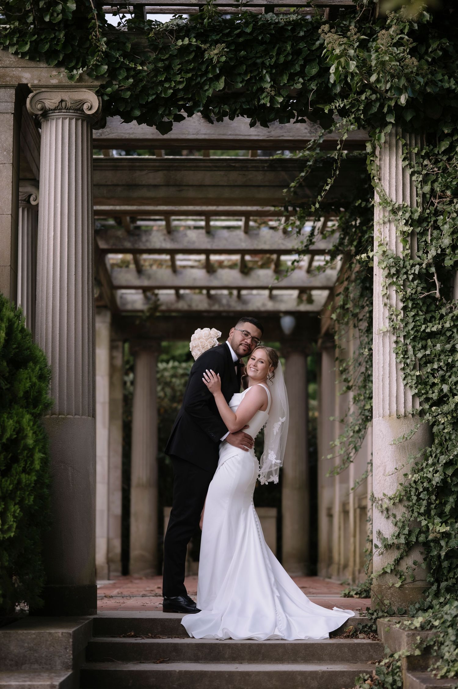 Wedding couple poses romantically under classical stone columns and vines at elegant venue.