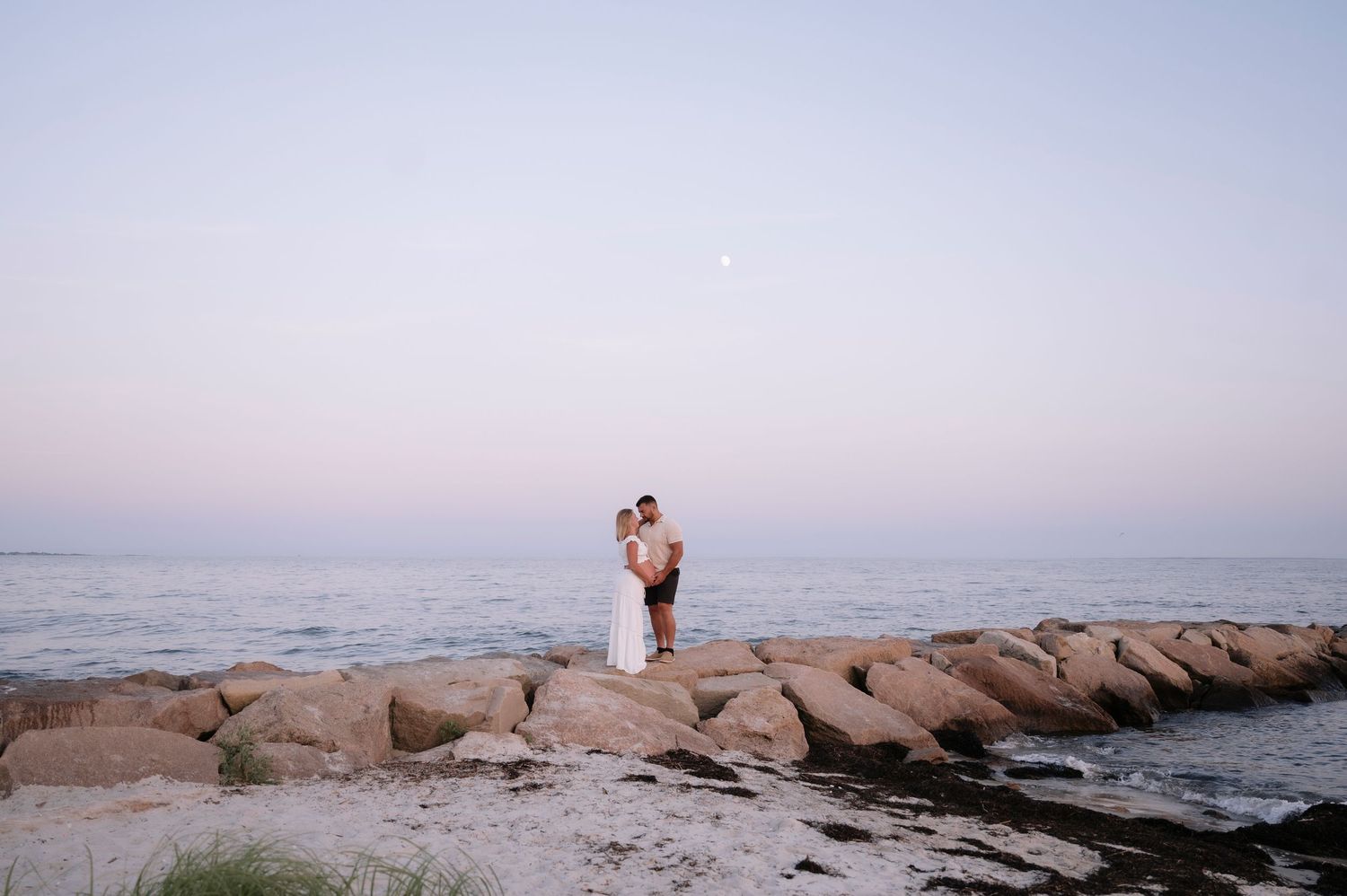 Couple embracing on rocky shoreline at dusk with calm ocean and pastel sky backdrop.