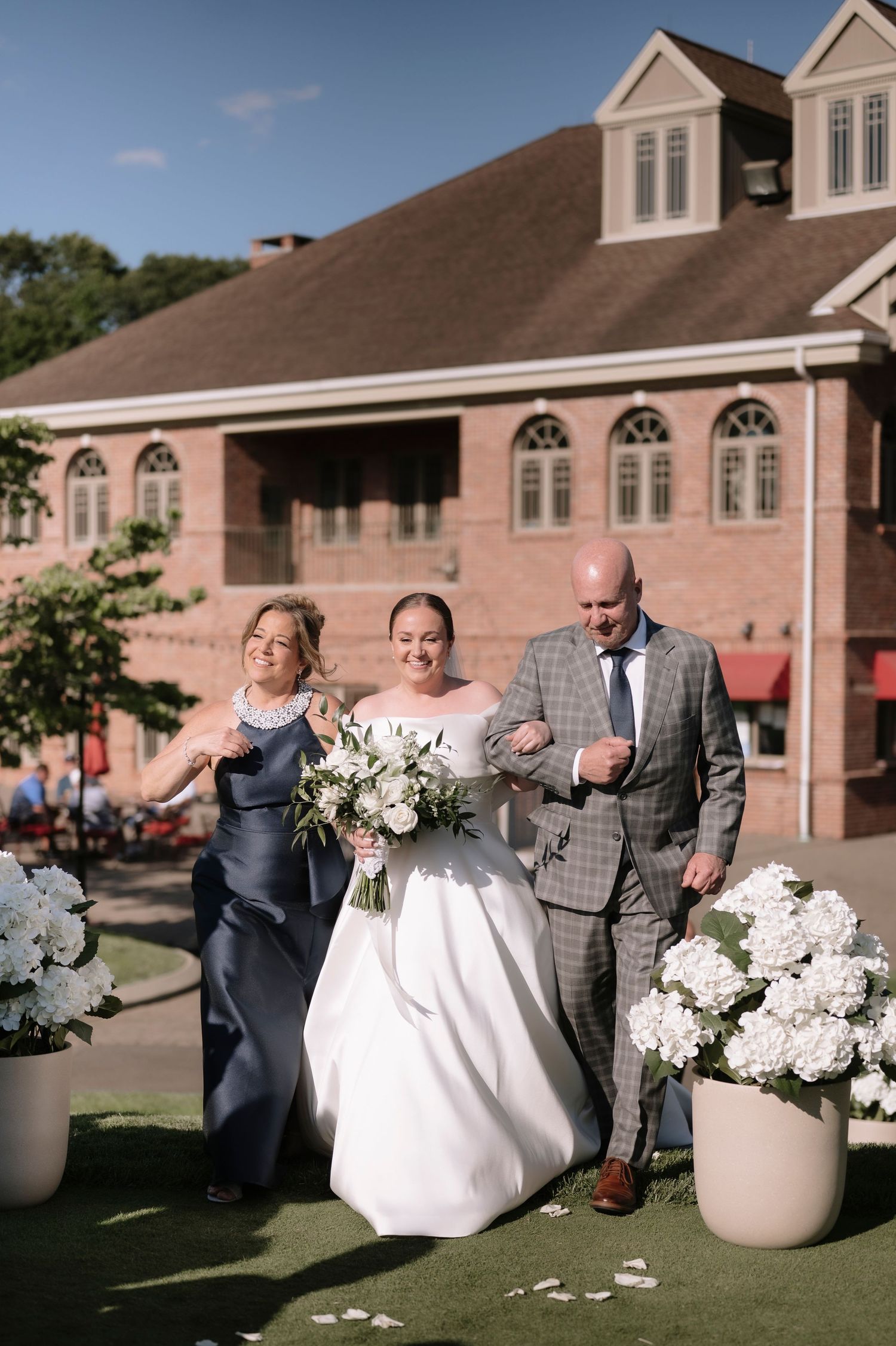 Bride walks down outdoor aisle with parents, holding white bouquet, beside hydrangea arrangements at brick building venue.