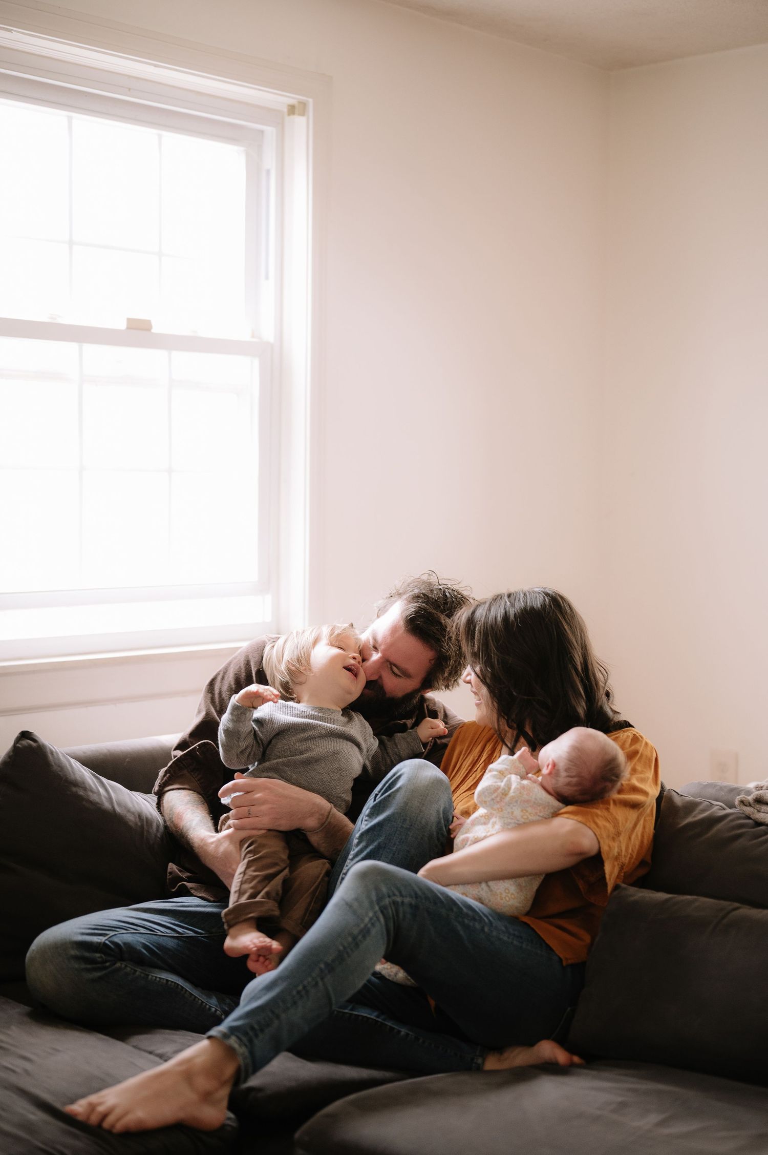 Family snuggling close together on a dark couch beneath a bright window.