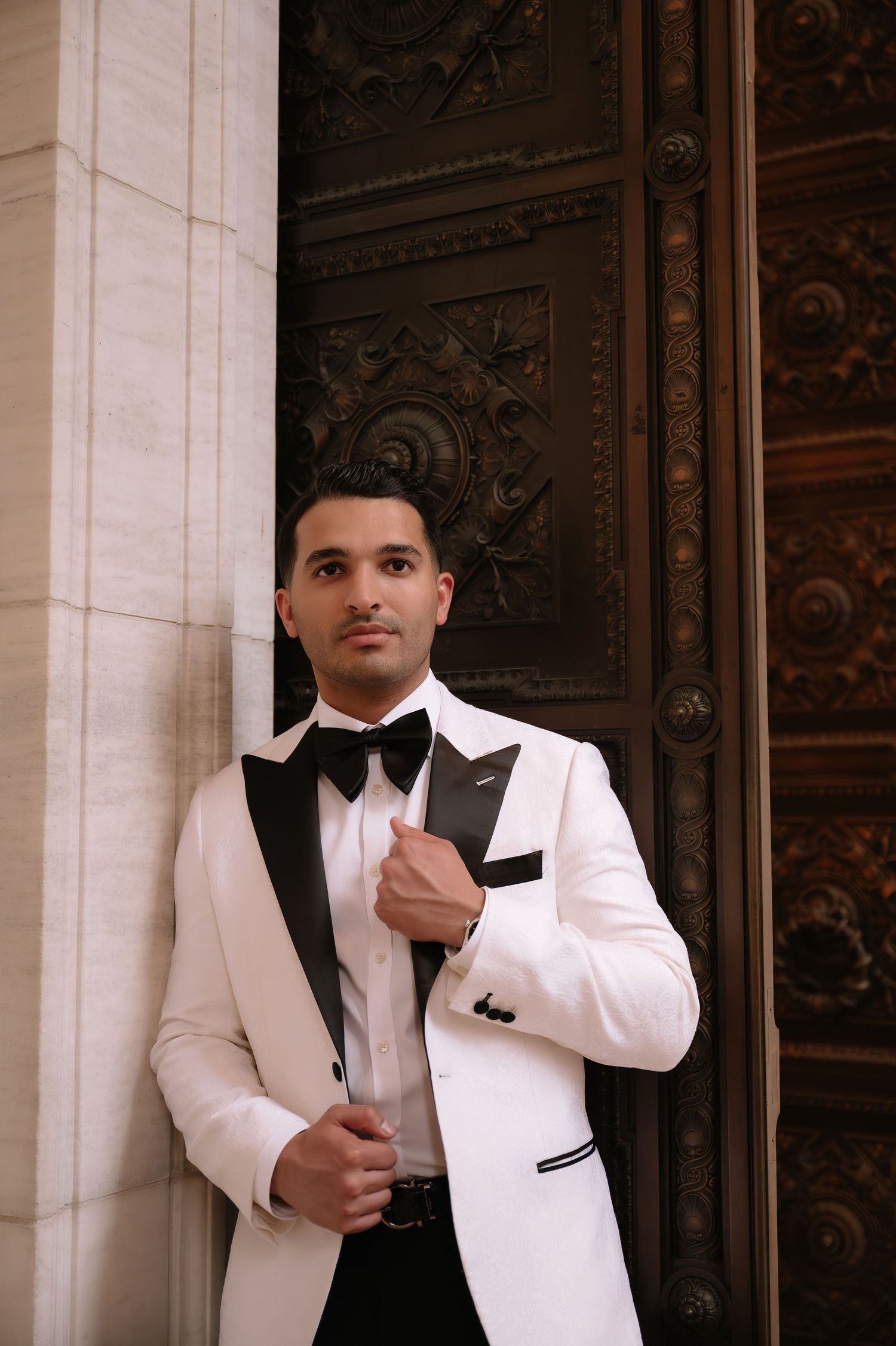 A person wearing a white tuxedo jacket with black lapels, black bow tie and matching accessories poses in front of ornate doors.