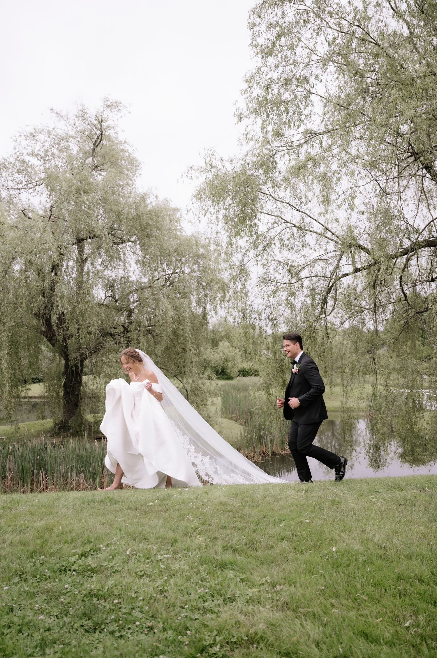 A couple in wedding attire sits on a grassy hillside under willow trees while a person in black stands nearby.
