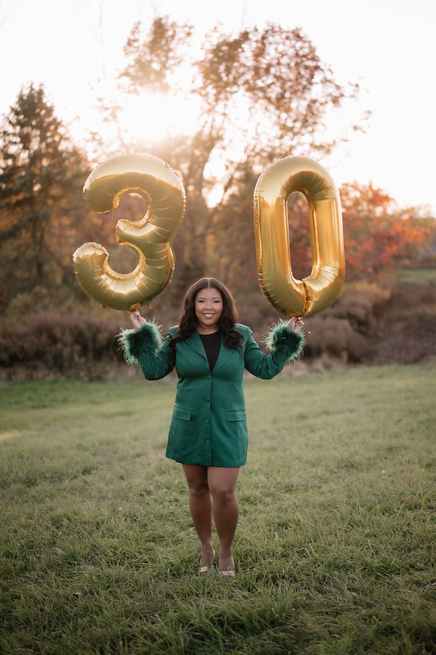 A person in a green winter coat holds up gold number 30 balloons in a grassy field during sunset.