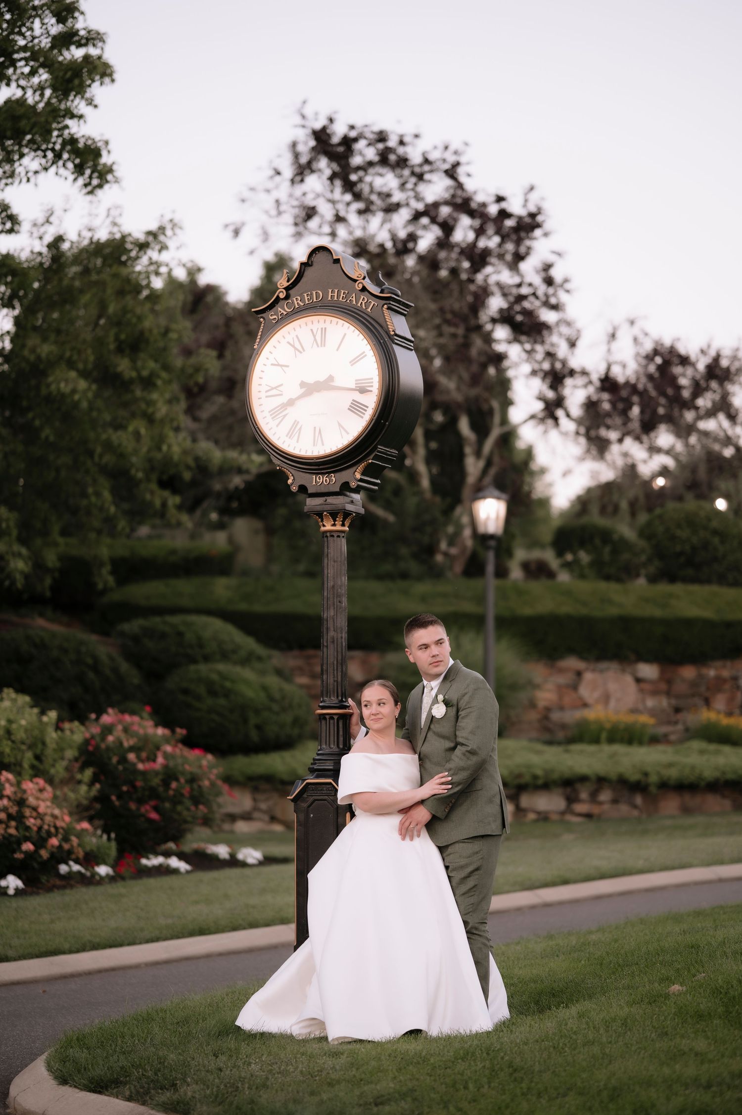 Bride and groom posing together beside an ornate outdoor clock at dusk in a lush garden setting.